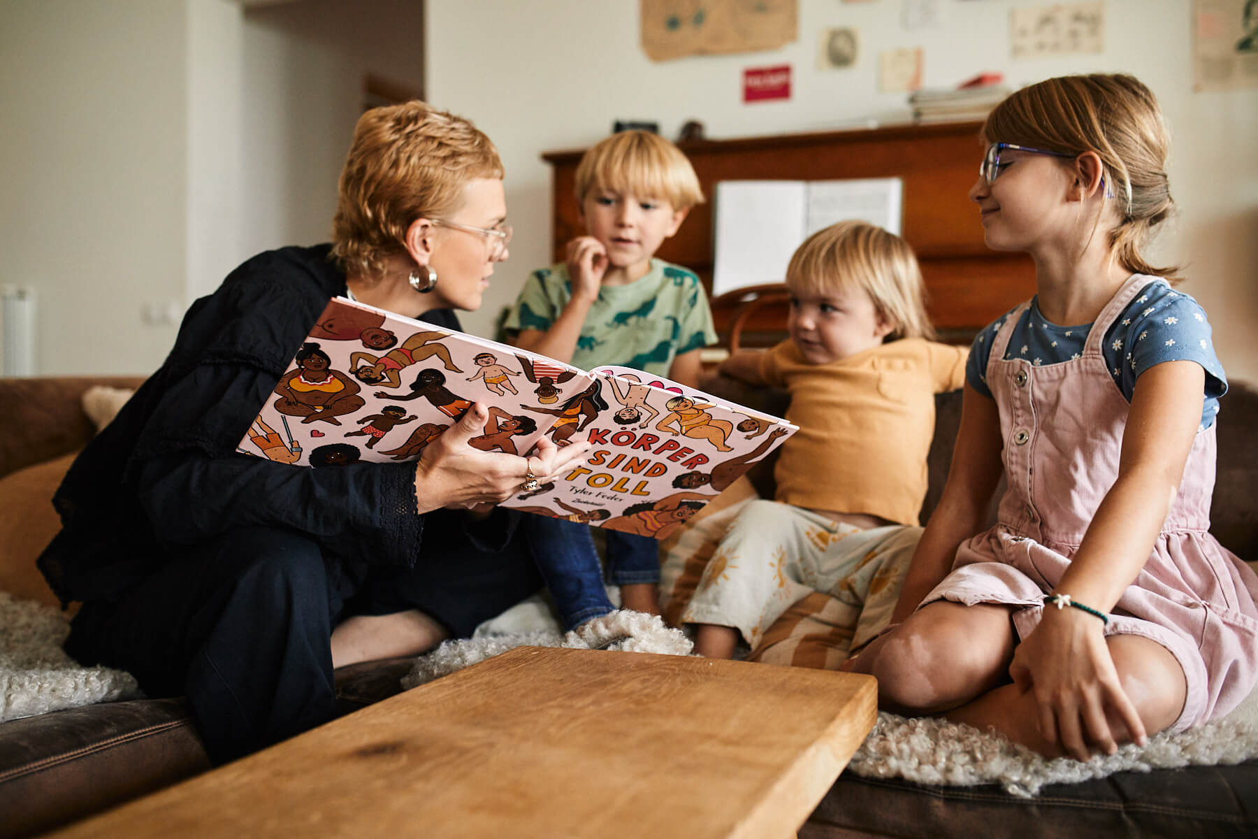 Ein Erwachsener liest drei kleinen Kindern, die auf der Couch sitzen, ein buntes Bilderbuch vor, und alle hören in einem gemütlichen Wohnzimmer aufmerksam zu.