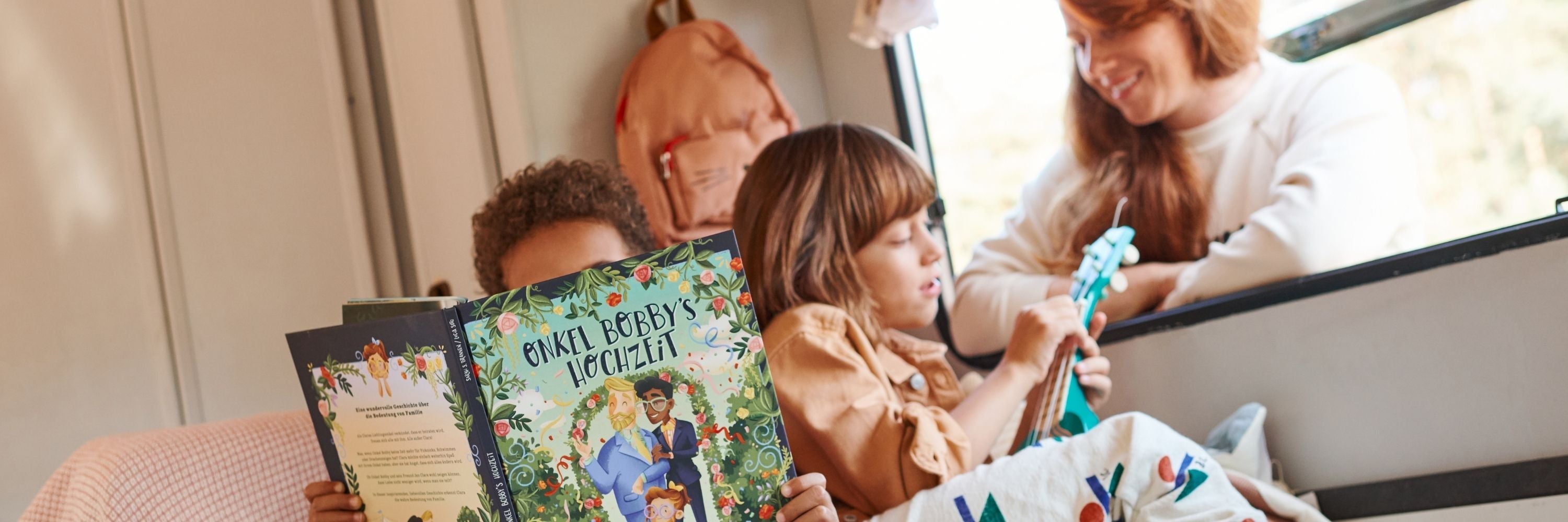 Zwei Kinder sitzen auf einem Sofa; eines liest ein buntes Buch mit dem Titel "Onkel Bobbys Hochzeit", während das andere eine kleine blaue Gitarre spielt. Eine Erwachsene lächelt ihnen durch ein nahes Fenster zu.