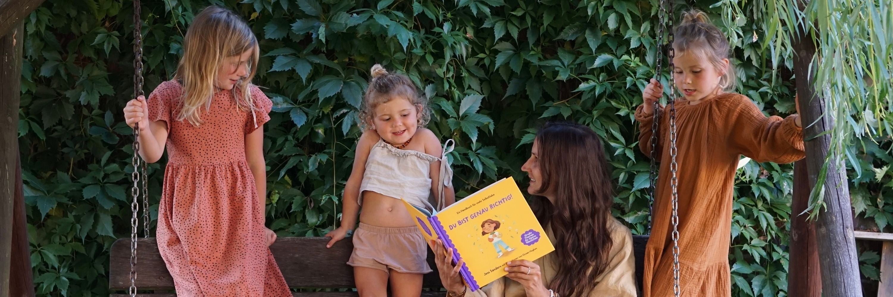 Eine Frau liest drei jungen Mädchen auf einer hölzernen Schaukelbank im Freien ein Kinderbuch vor, mit grünen Blattpflanzen im Hintergrund. Zwei Mädchen stehen auf der Schaukel, und eines sitzt auf der Bank.