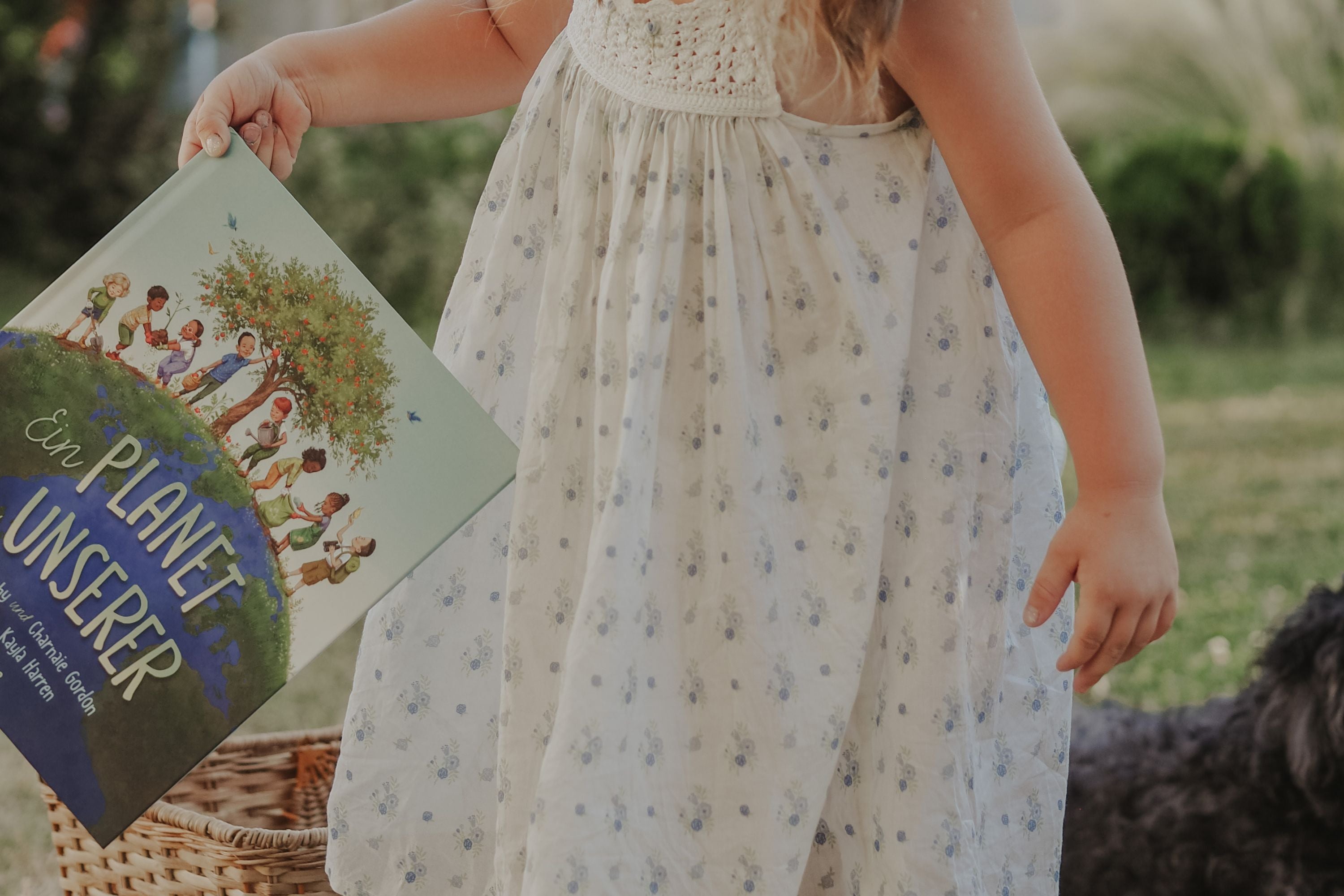 Ein kleines Kind in einem weißen geblümten Kleid hält ein Buch mit dem Titel Ein Planet für uns im Freien in der Nähe eines Weidenkorbs und eines schwarzen Hundes, mit grünem Gras und Sträuchern im Hintergrund.