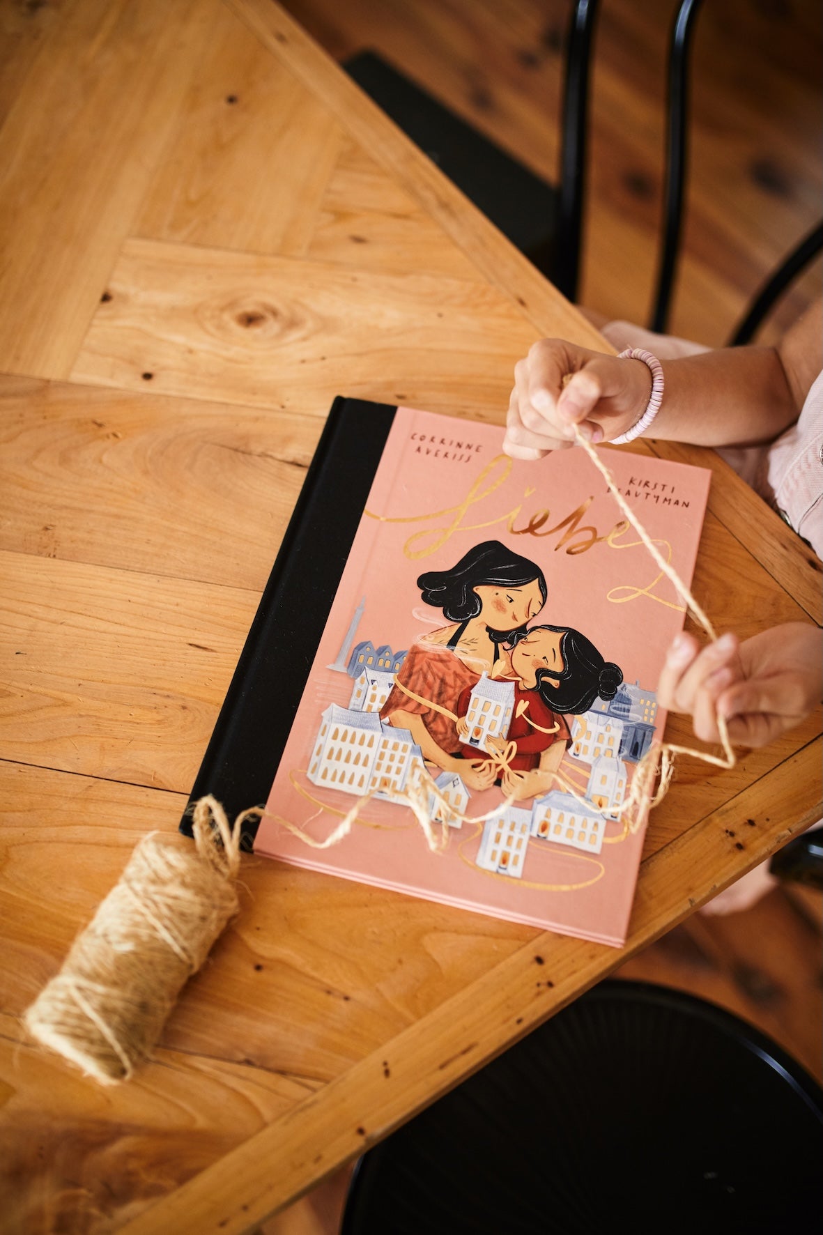Ein Kind sitzt an einem Holztisch und hält eine Schnur neben dem Buch Das Kennlern-Paket des Zuckersüß Verlags, das ein illustriertes Cover mit einer Frau zeigt, die ein Kind umarmt, während im Hintergrund Gebäude zu sehen sind. Das Buch liegt neben einer Schnurspule.