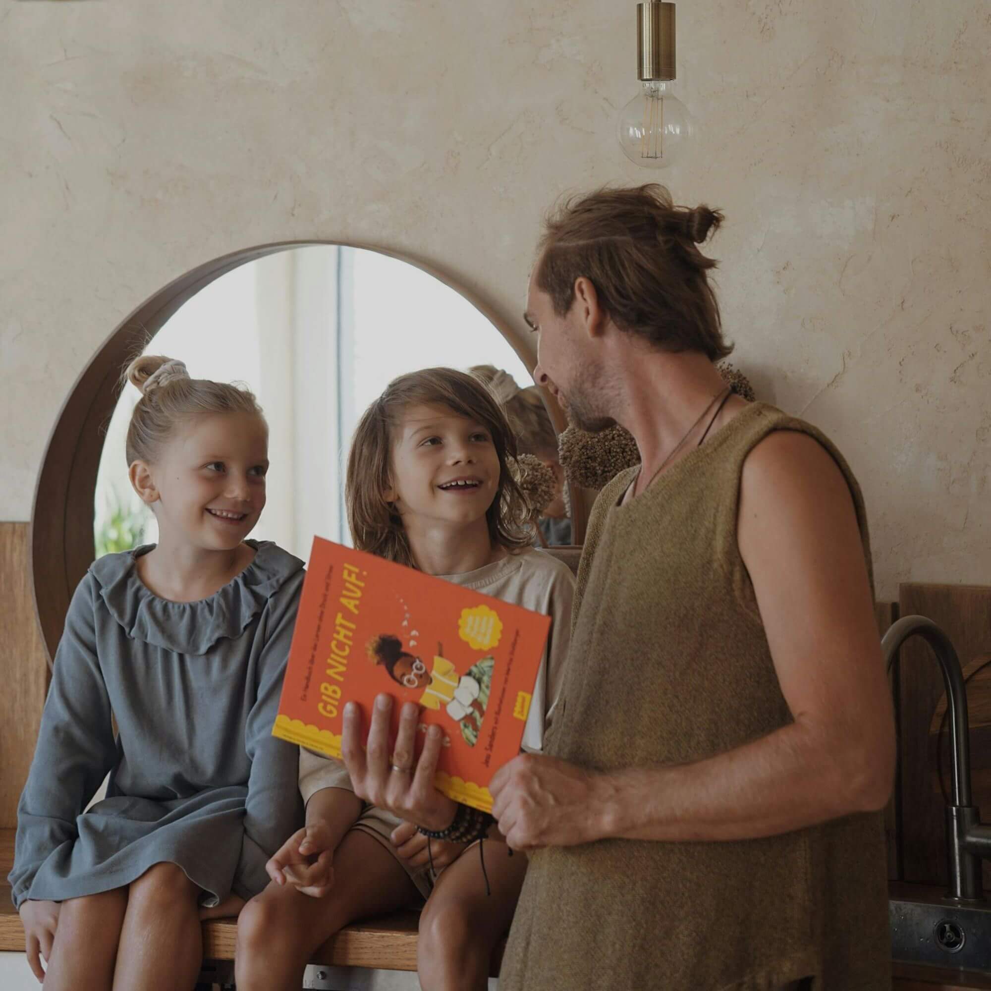 Ein Erwachsener hält "Gib nicht auf!" von Jessica Sanders in der Hand und setzt sich mit zwei lächelnden Kindern vor einen runden Spiegel. Die Kinder hören aufmerksam zu und genießen den gemütlichen Moment.