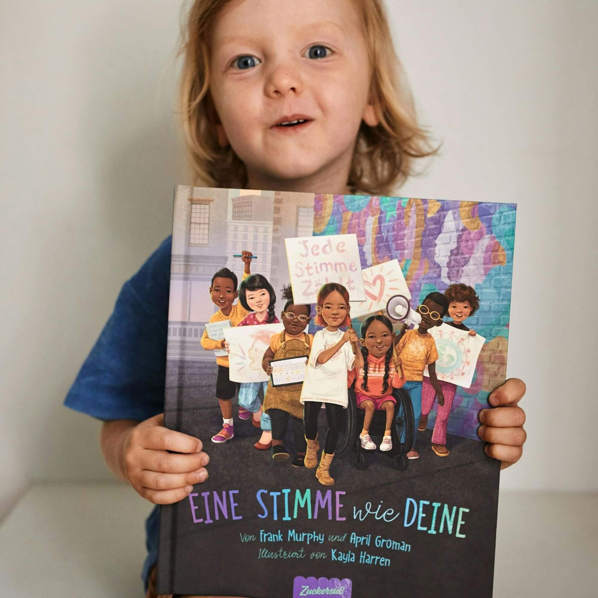 Ein kleines Kind mit hellem Haar und blauem Hemd hält "Eine Stimme wie deine" von Frank Murphy und April Groman in die Höhe. Auf dem Cover sind verschiedene Kinder zu sehen, die Schilder mit Sätzen über Stimme und Kommunikation halten.