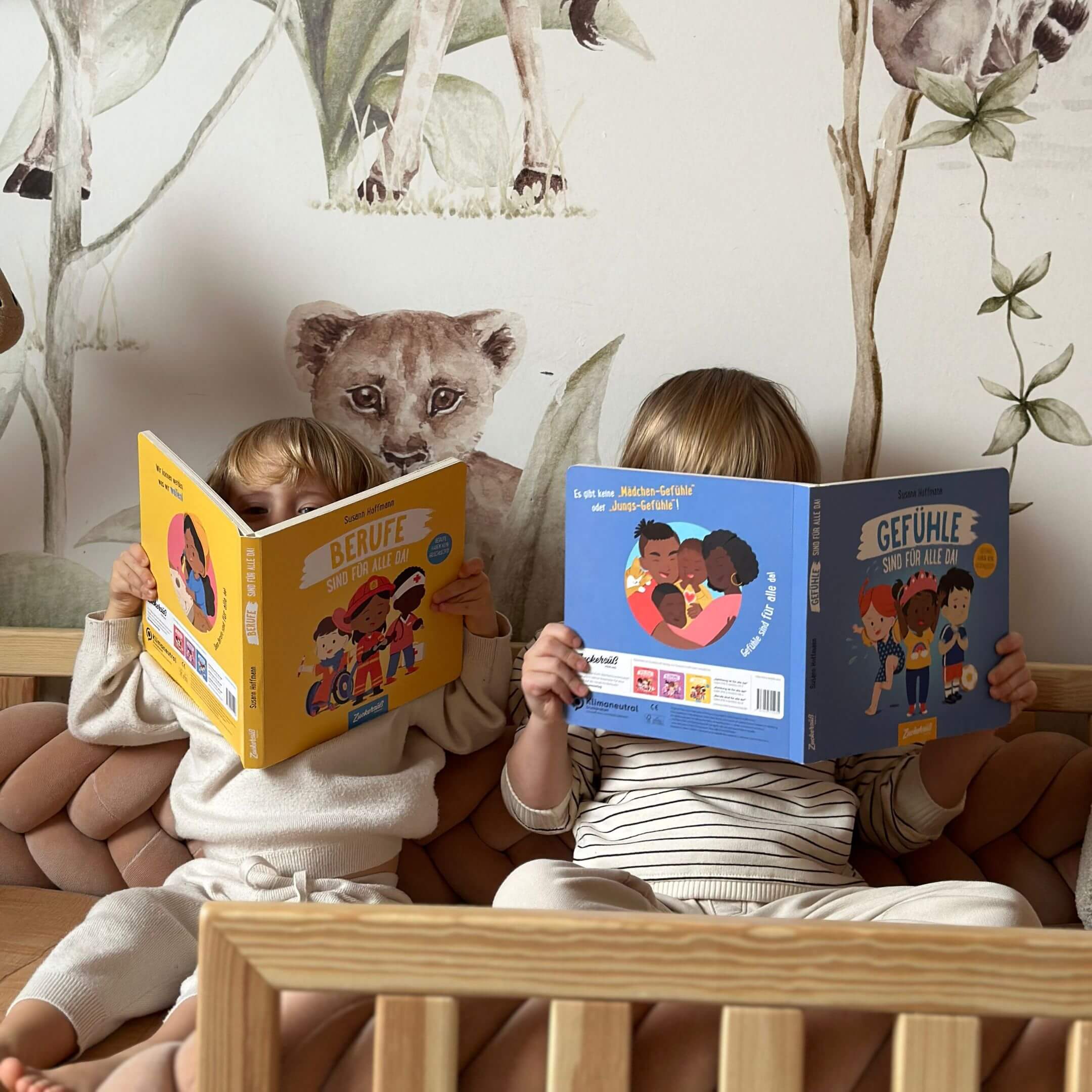 Zwei Kinder sitzen nebeneinander auf einer Couch, halten Susann Hoffmanns "Berufe sind für alle da" & "Gefühle sind für alle da" in der Hand und lesen, ihre Gesichter hinter den Büchern verborgen. Tierillustrationen, darunter ein Löwenjunges, schmücken die Wand hinter ihnen.
