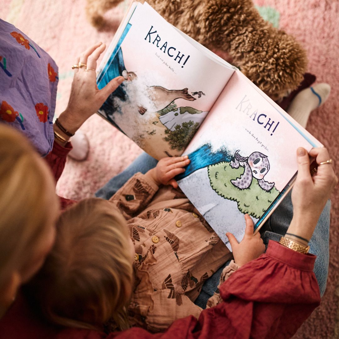 Ein Erwachsener und ein Kind lesen Nellies großen Sprung vom Zuckersüß Verlag, ein illustriertes Buch über Mut und innere Stärke. Das Buch liegt aufgeschlagen auf ihrem Schoß, Tierzeichnungen sind zu sehen, und ein Plüschteddy sitzt in der Nähe auf dem rosa Teppich.