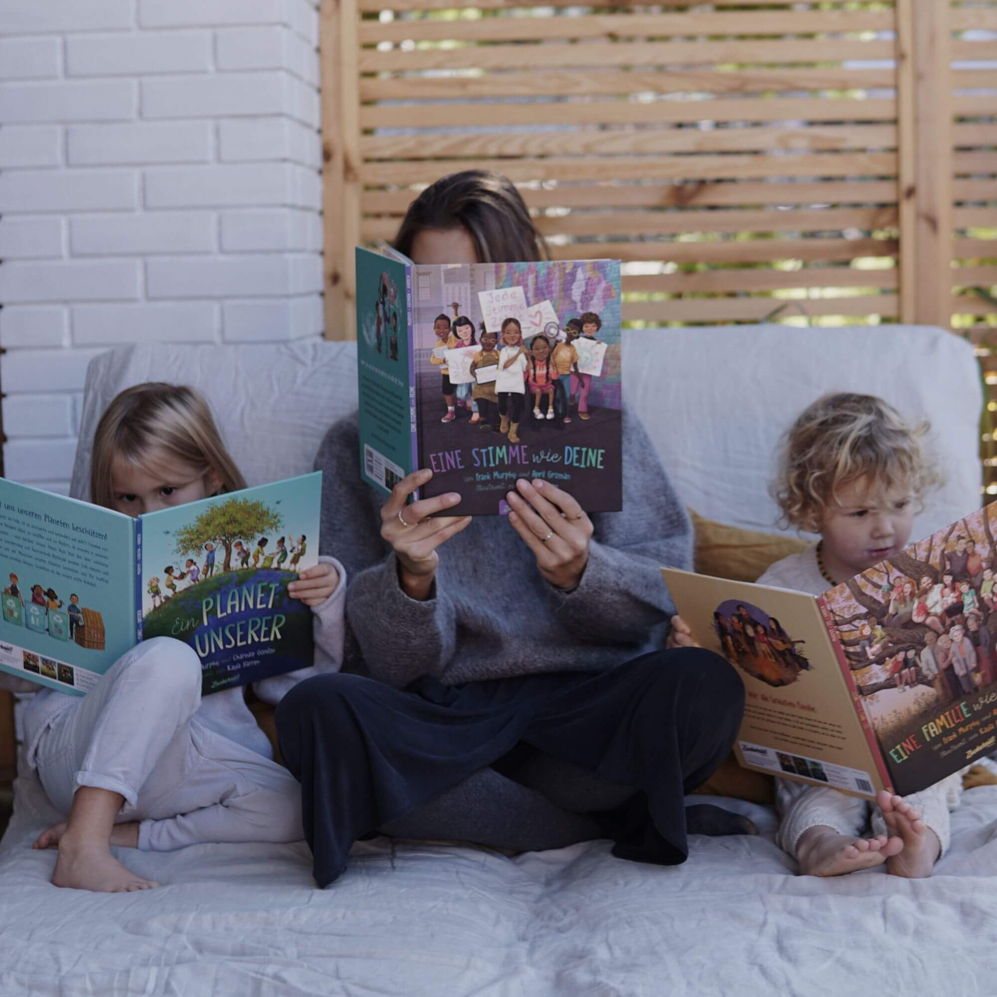 Ein Erwachsener und zwei Kinder lesen gemeinsam auf einer gemütlichen Couch, jeder mit einem aufgeschlagenen Buch. Das "Welt verändern" Starter Pack aus dem Zuckersüß Verlag inspiriert Kinder und vermittelt wichtige Werte in entspannter Atmosphäre.