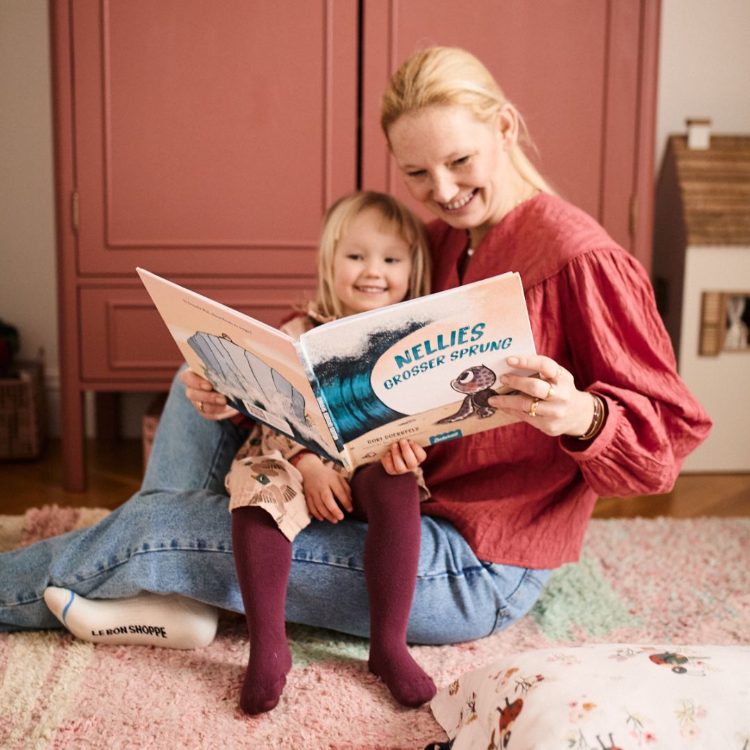 Eine lächelnde Frau und ein kleines Mädchen sitzen auf dem Boden und lesen "Nellies großer Sprung" aus dem Zuckersüß Verlag, ein Kinderbuch über Mut und innere Stärke, in einem gemütlichen Zimmer mit einem rosa Schrank und einem Puppenhaus im Hintergrund.