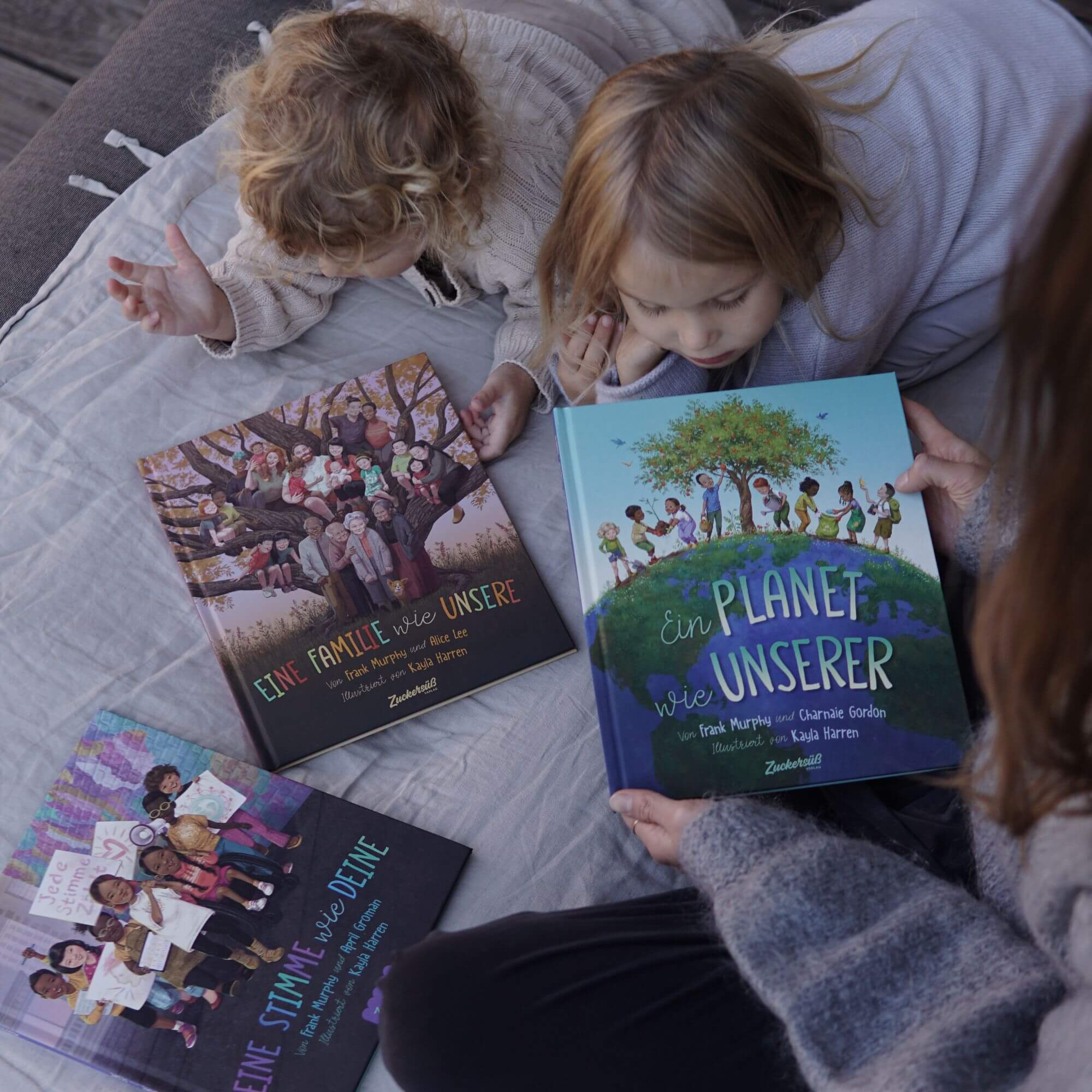 Zwei Kinder lesen Bilderbücher aus dem "Welt verändern" Starter Pack des Zuckersüß Verlags. Ein Erwachsener hält eines auf; drei bunte Cover zeigen unterschiedliche Menschen, die wichtige Werte vermitteln und Kinder dazu inspirieren, etwas zu bewegen.
