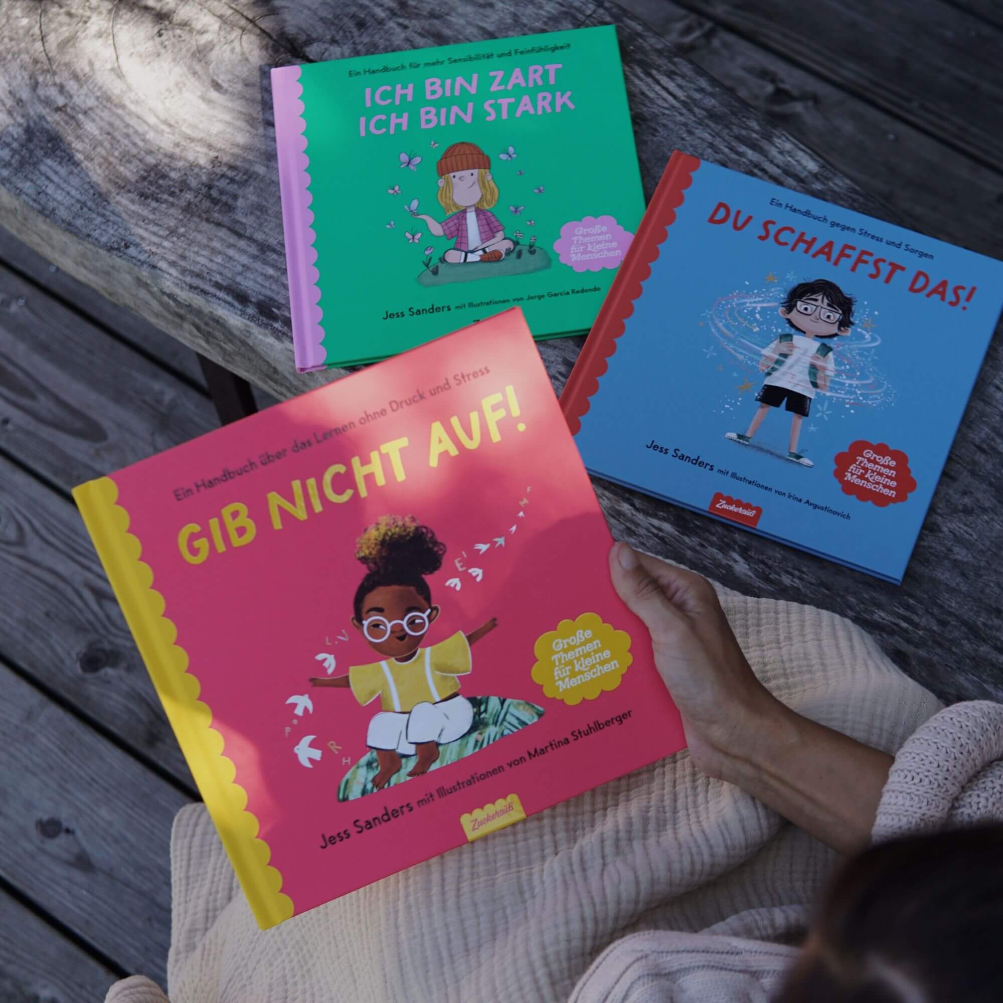 Drei farbenfrohe Kinderbücher von Jess Sanders aus dem Mutmach-Paket des Zuckersüß Verlags liegen auf einer Holzbank. Eine Person hält das Buch „Gib nicht auf!“ in der Hand, daneben liegen „Ich bin zart, ich bin stark“ und „Du schaffst das!“. Die Szene vermittelt Ruhe und lädt zum gemeinsamen Lesen über Gefühle, Stärke und Selbstvertrauen ein.