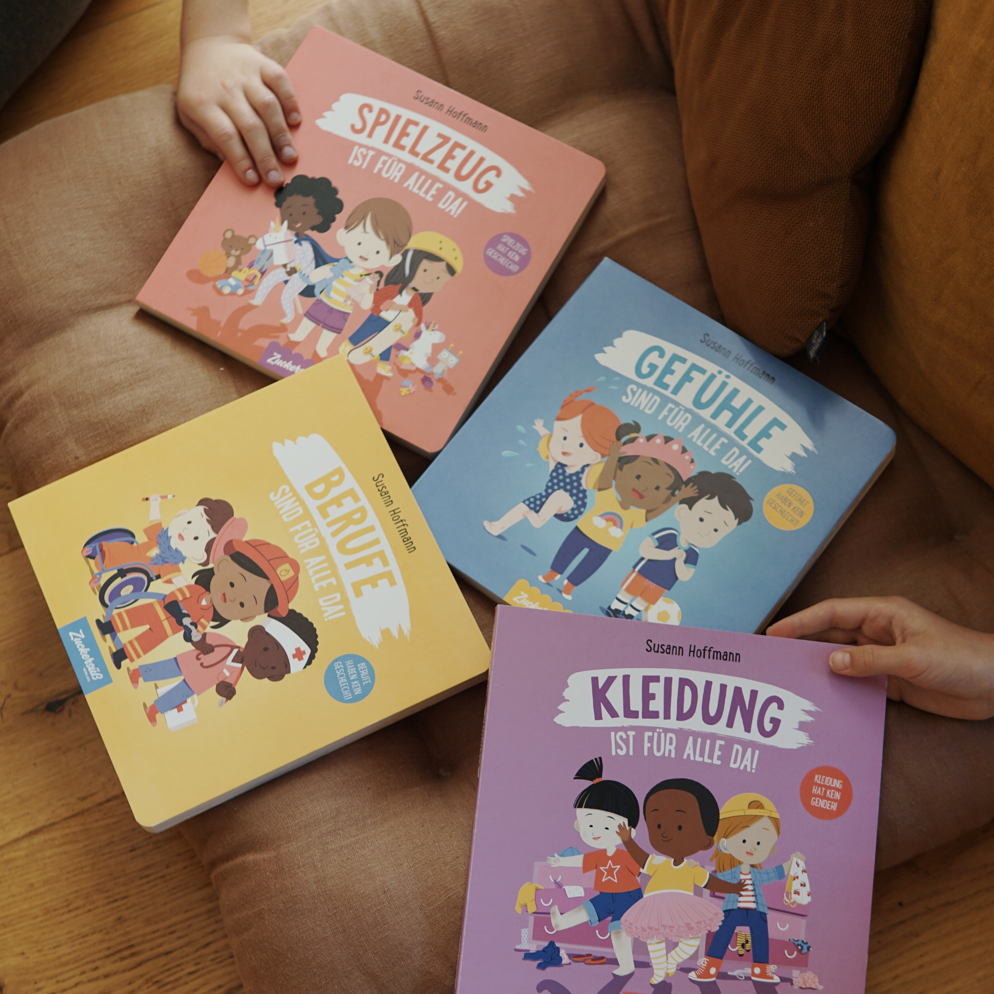 Vier illustrierte Kinderbücher von Susann Hoffmann aus der Reihe „Freiheit ist für alle da“ vom Zuckersüß Verlag liegen auf einem Holzboden neben einem braunen Sofa. Zwei Kinderhände greifen nach den Büchern. Die farbenfrohen Cover behandeln Vielfalt und Gleichberechtigung in den Bereichen Spielzeug, Gefühle, Berufe und Kleidung.