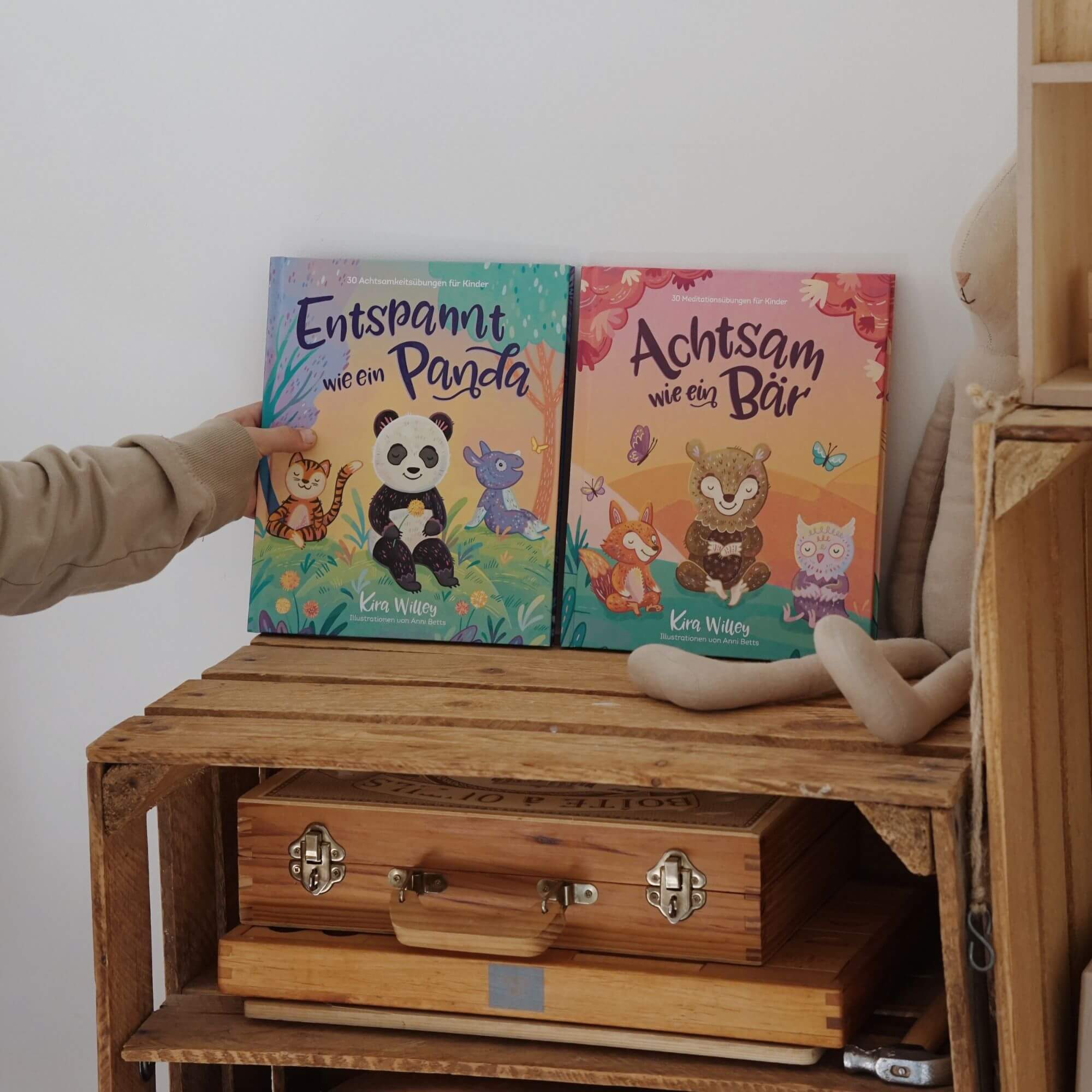 Eine Hand hält "Entspannt wie ein Panda" neben "Achtsam wie ein Bär" aus dem "Achtsamkeit im Doppelpack" des Zuckersüß Verlags. Die beiden Kinderbücher mit Meditationsübungen liegen auf einem Holzregal mit Spielzeug und Kiste.