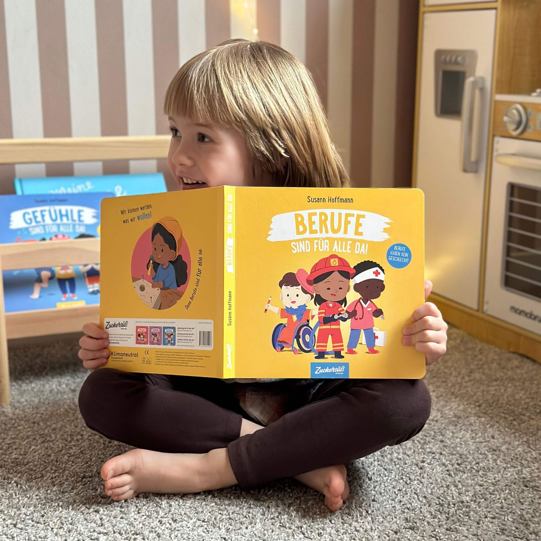 Ein kleines Kind mit hellbraunem Haar sitzt im Schneidersitz auf einem Teppich, lächelt und hält das Susann Hoffmann Pappbilderbuch "Berufe sind für alle da!" in der Hand, das auf dem Umschlag verschiedene Figuren zeigt, die als unterschiedliche Berufe verkleidet sind.