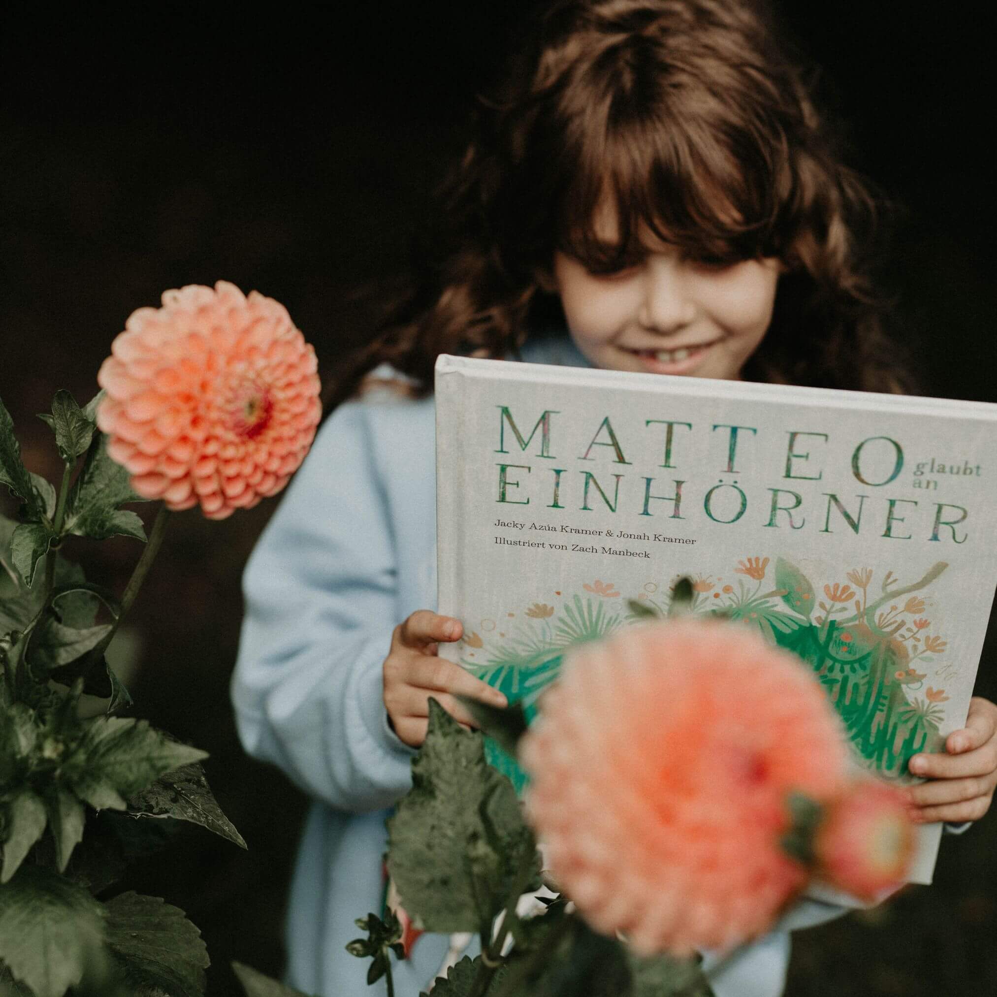 Ein kleines Kind mit lockigen braunen Haaren in einem hellblauen Pullover lächelt, während es "Matteo glaubt an Einhörner" von Jackie Azúa Kramer & Jonah Kramer in der Nähe blühender rosa Blumen im Freien liest.