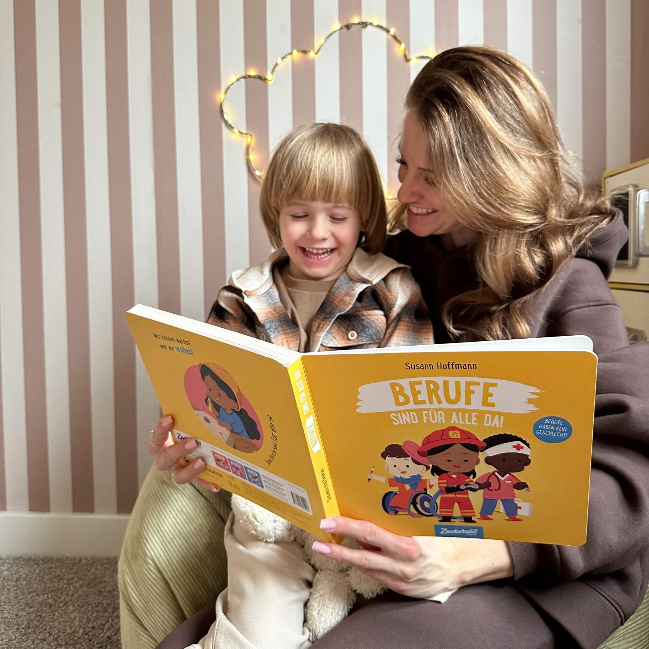 Eine lächelnde Frau und ein Kind sitzen zusammen auf einem Sitzsack und lesen "Berufe sind für alle da!" von Susann Hoffmann. Gestreifte Tapeten und eine herzförmige Lichterkette schaffen eine gemütliche Atmosphäre.