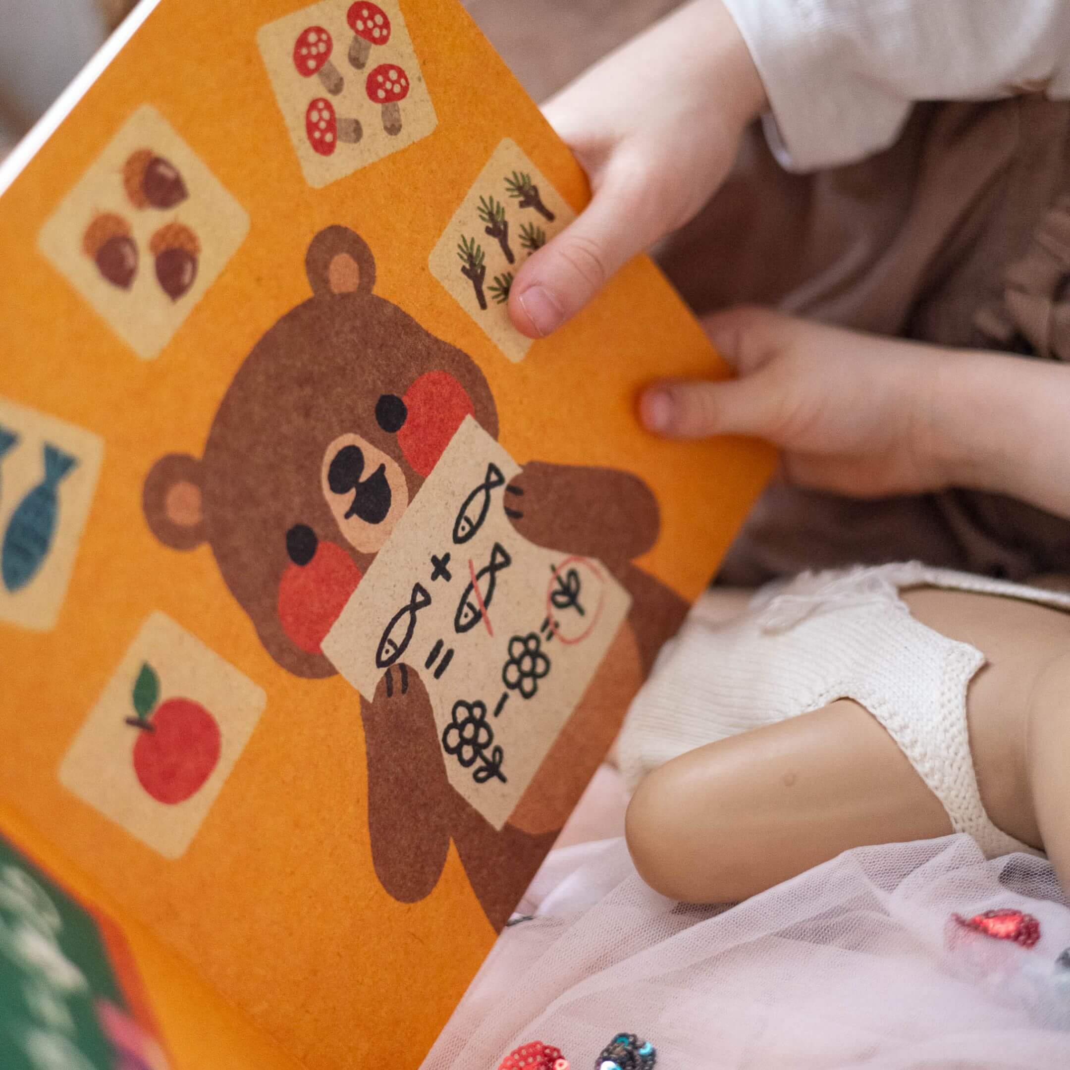 Ein Kind hält "Hilfe brauchen ist okay" & "Fehler machen ist okay" von Annelies Draws, das einen illustrierten Braunbär mit Pilzen, Kräutern, Fisch und einer Tomate zeigt. Die Hände des Kindes und eine Puppe im weißen Kleid sind teilweise sichtbar.