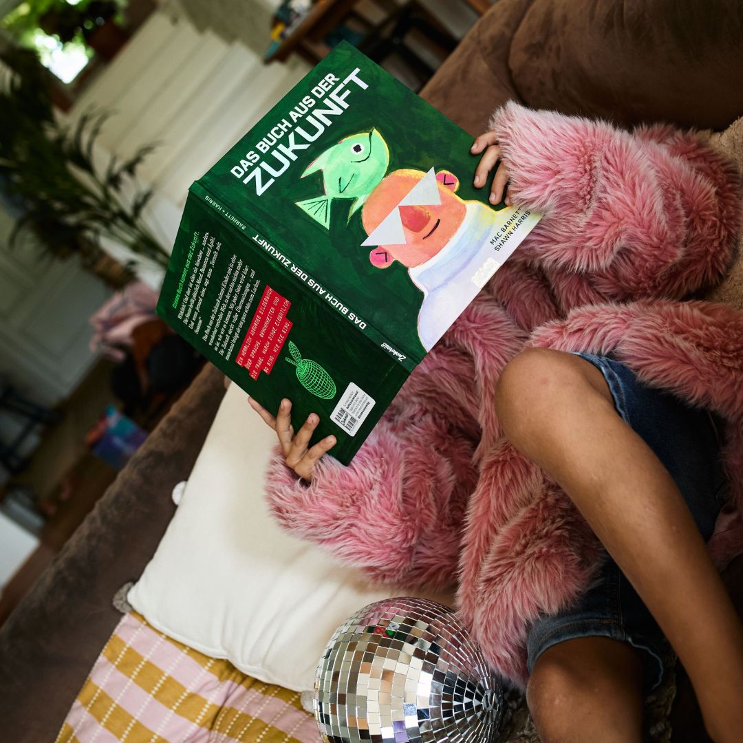 Eine Person in einer rosa Pelzjacke entspannt sich auf einem Sofa und liest „Das Buch aus der Zukunft“ vom Zuckersüß-Verlag, neben ihr steht eine Discokugel und im Hintergrund ist eine Pflanze zu sehen.