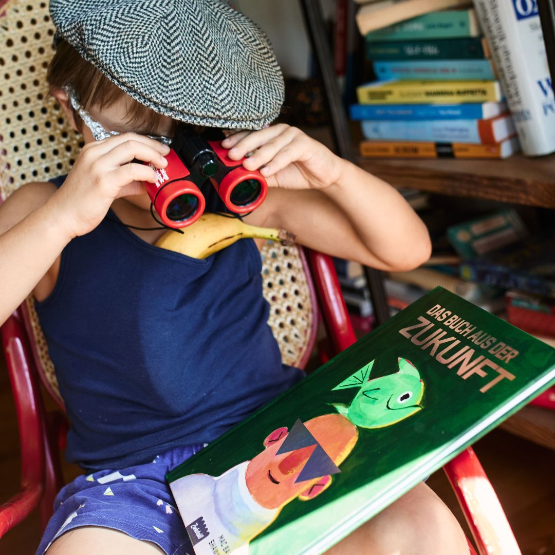 Ein Kind mit einer Mütze auf dem Kopf und einem roten Fernglas in der Hand sitzt mit einer Banane um den Hals neben dem deutschen Buch „Das Buch aus der Zukunft“ vom Zuckersüß-Verlag. Im Hintergrund sind Bücherstapel zu sehen.