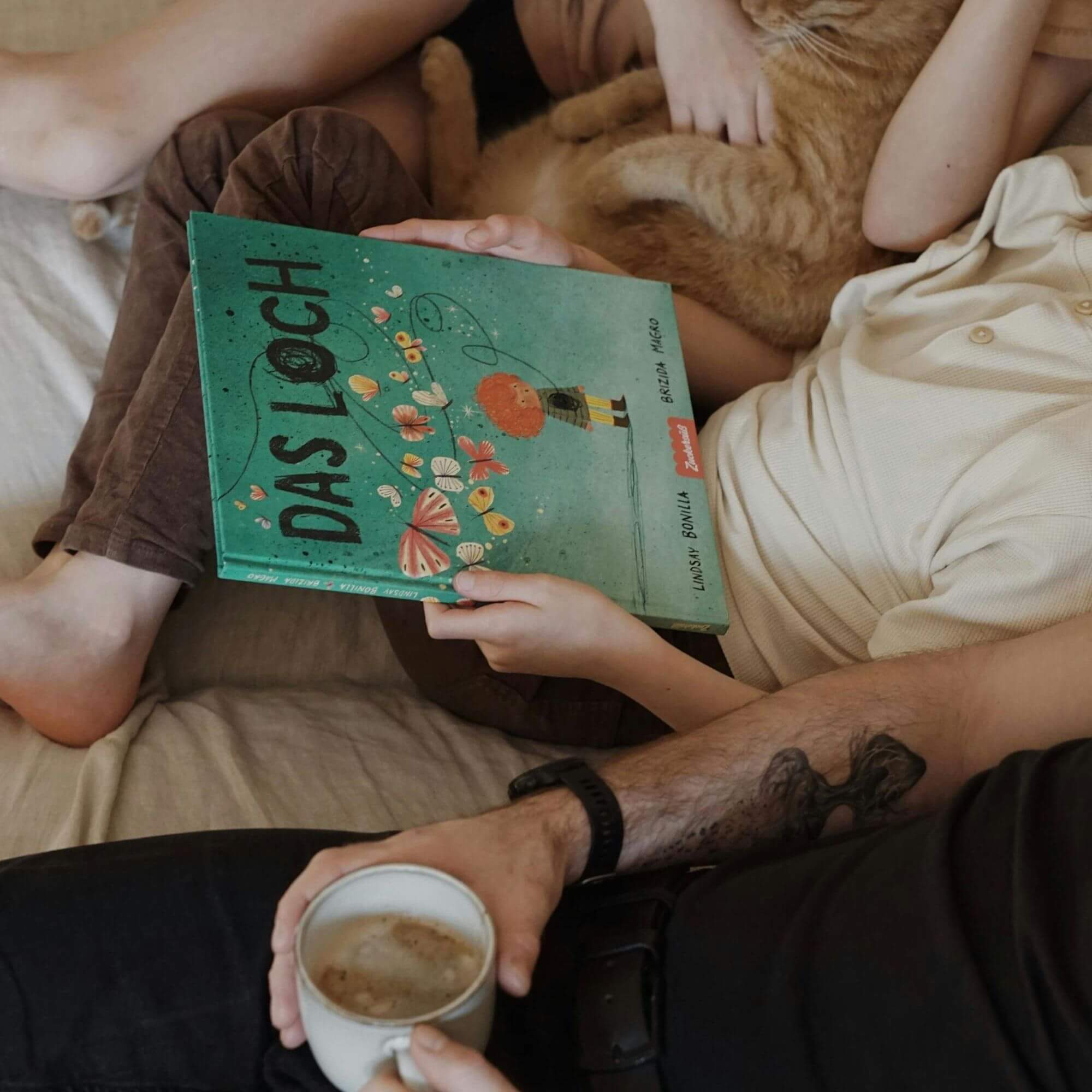 Eine gemütliche Szene: Jemand hält das grüne Buch "Das Loch" von Lindsay Bonilla in der Hand, während er mit einem Kind und einer orangefarbenen Katze auf einem Bett liegt; eine andere Person in der Nähe genießt einen Kaffee und schafft so eine entspannte Atmosphäre der Erinnerung und des sanften Heilens.