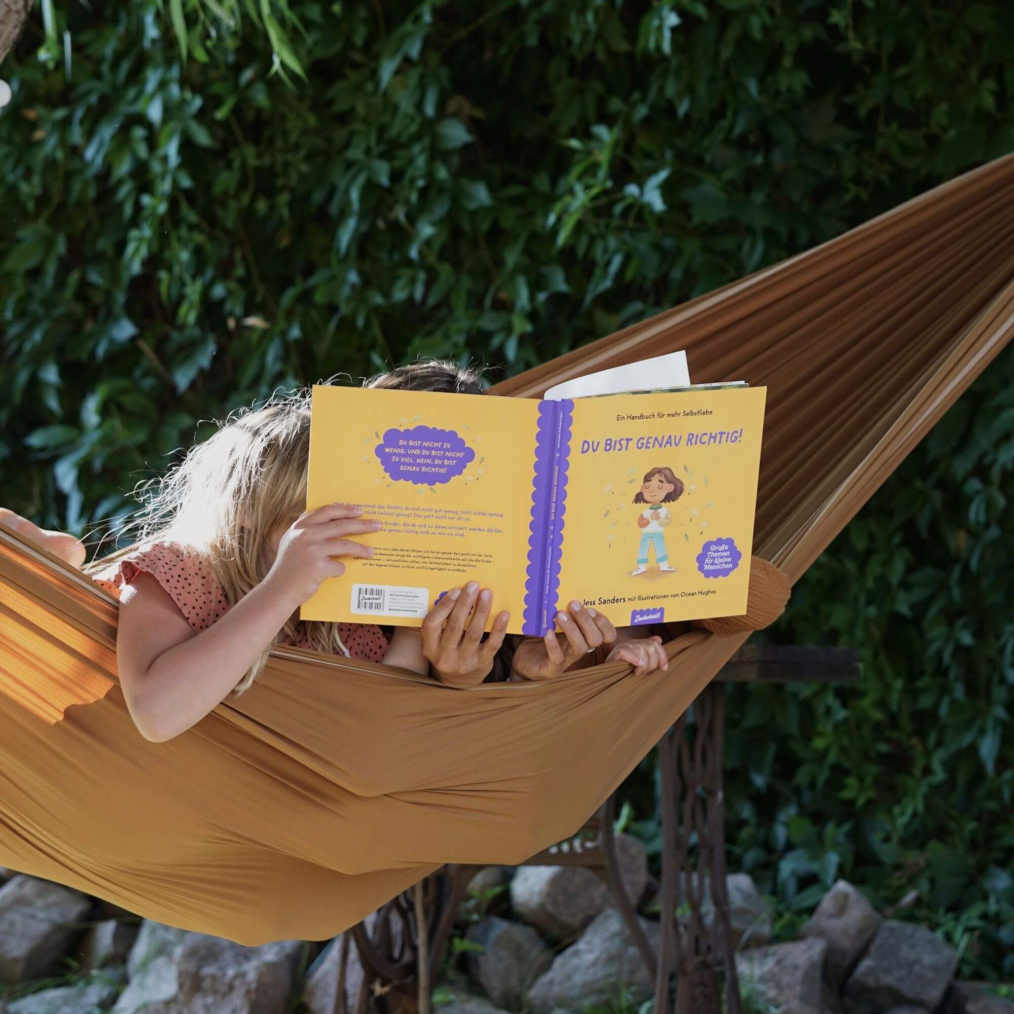 Zwei Kinder entspannen sich gemeinsam in einer braunen Hängematte im Freien und lesen