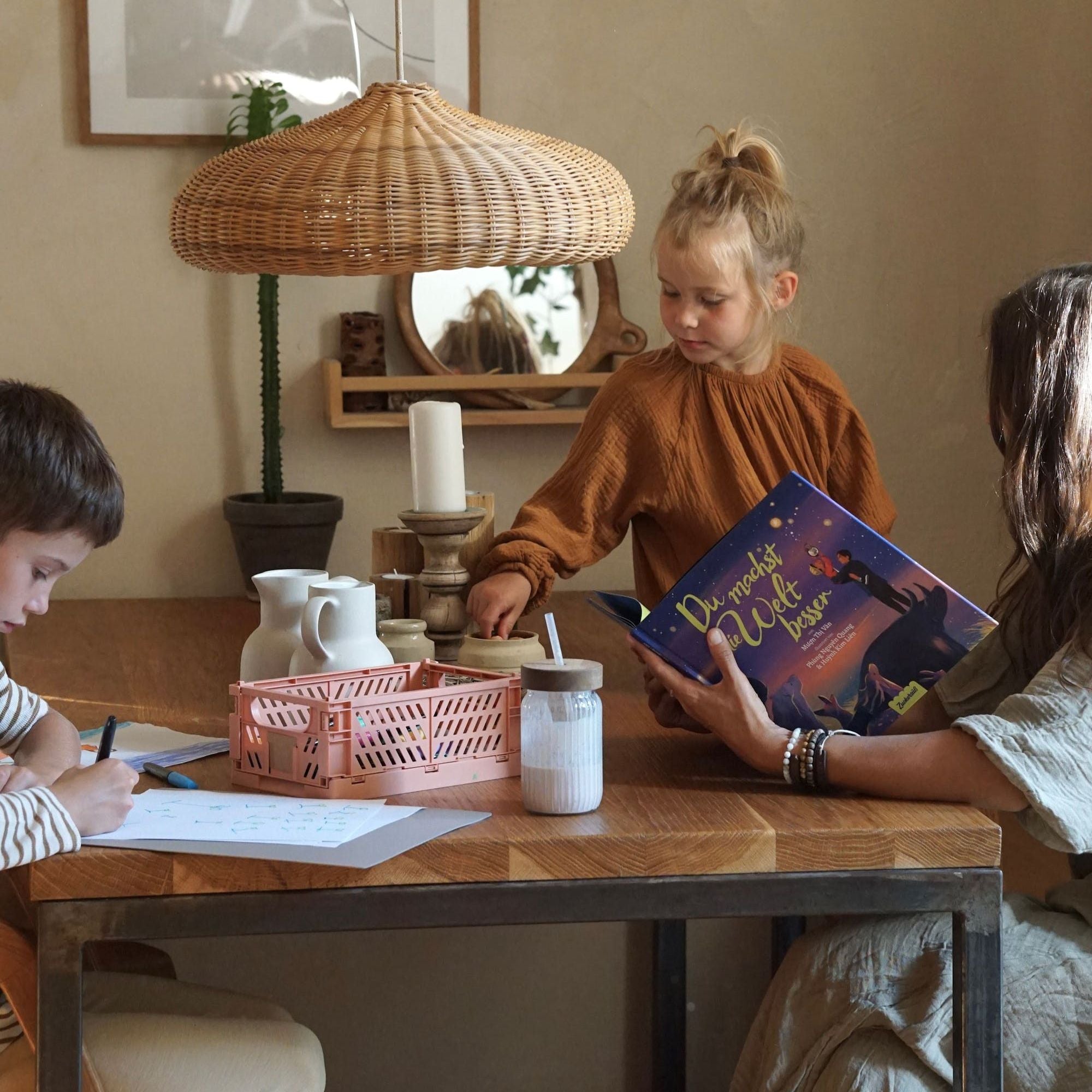 Eine Frau und zwei Kinder sitzen an einem Holztisch; ein Kind zeichnet, während das andere hinübergreift. Die Frau hält