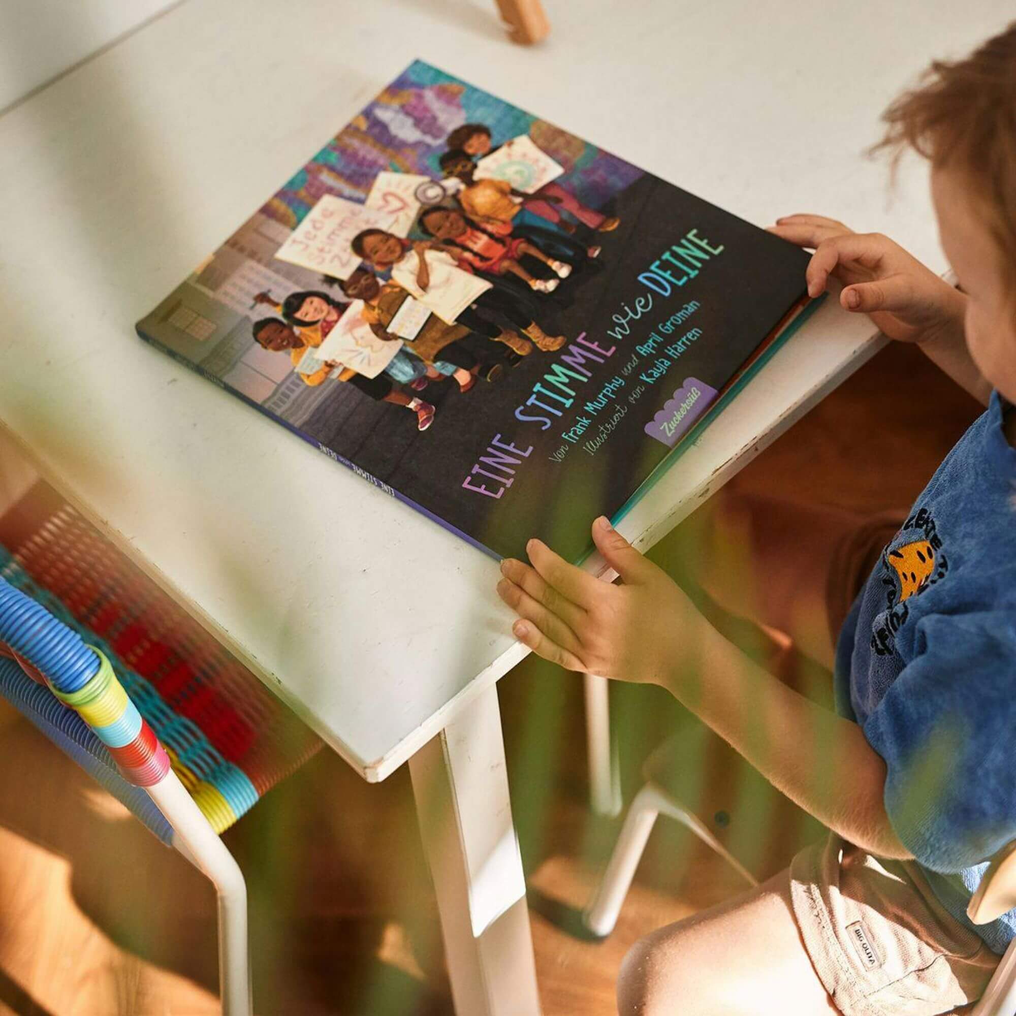 Ein Kind sitzt an einem weißen Tisch und hält das Buch „Eine Stimme wie deine“. Auf dem Cover sind mehrere illustrierte Kinder zu sehen, die Schilder in den Händen halten. Daneben steht ein bunter Stuhl.