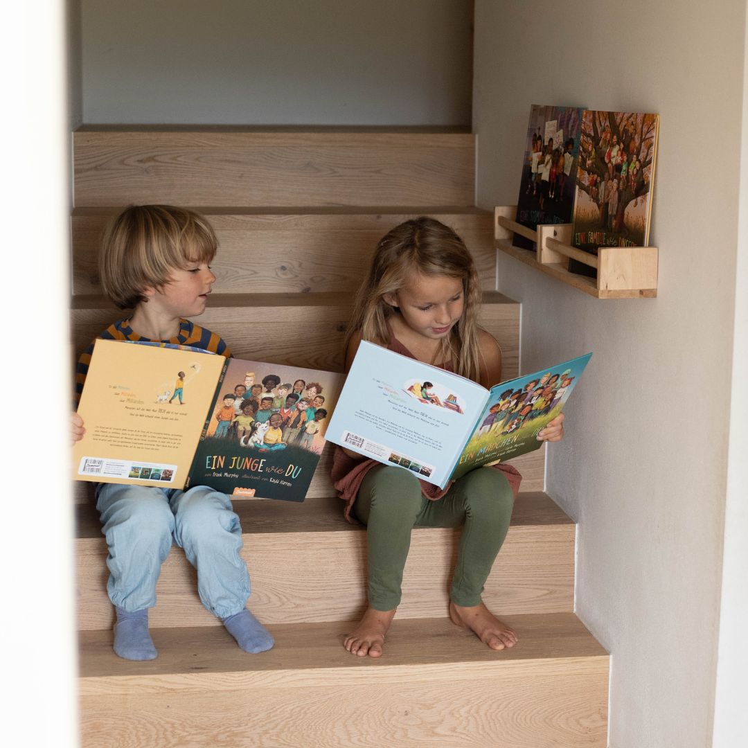 Zwei Kinder sitzen auf einer Holztreppe und lesen jeweils ein Bilderbuch aus der "Eine Welt wie diese - Mädchen-Kollektion" des Zuckersüß Verlags. In einem Wandregal darüber sind weitere Titel aus der Sammlung ausgestellt, die eine ruhige und gemütliche Atmosphäre schaffen.