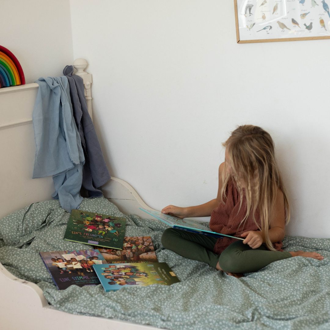 Ein kleines Kind mit langen blonden Haaren sitzt auf einem Bett mit grüner Bettwäsche und liest "Eine Welt wie diese - Mädchen-Kollektion" vom Zuckersüß Verlag. Andere Bücher aus der Reihe liegen in der Nähe; ein Regenbogenspielzeug und Kleidung hängen über dem Bett, und ein Vogelposter ziert die Wand.