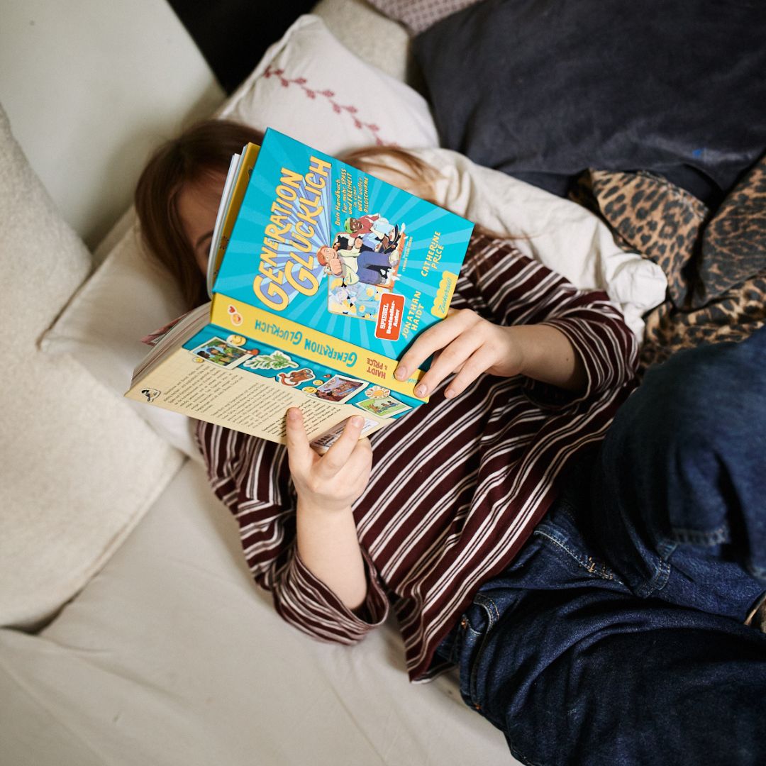 Ein Kind in gestreiftem Hemd und Jeans liegt auf einem Bett, das Gesicht hinter "Generation Glücklich" vom Zuckersüß Verlag verborgen. Umgeben von Kissen und Decken ziehen sie Geschichten der Bildschirmarbeit vor.