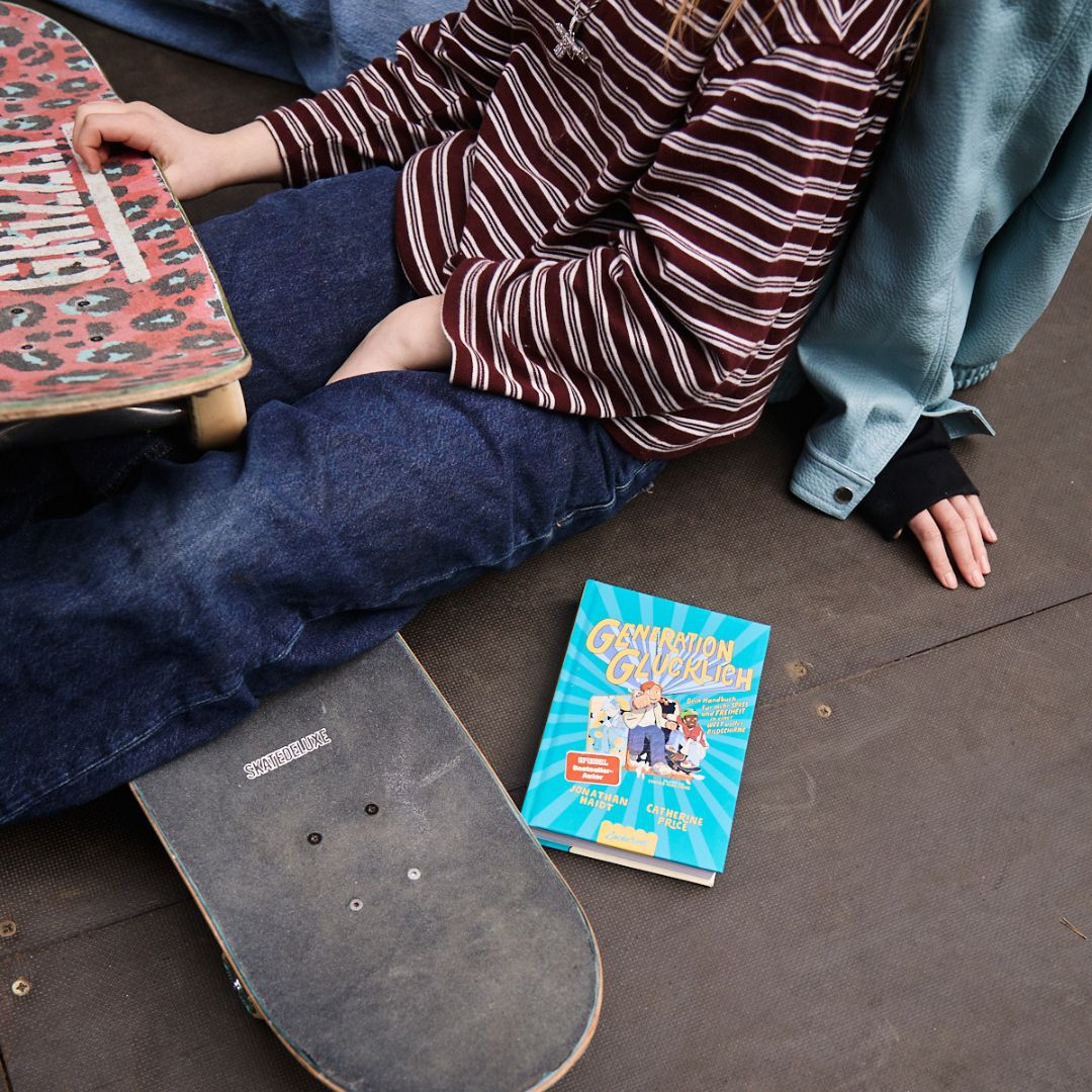Zwei Personen sitzen mit Skateboards auf dem Boden; eine hält ein Brett mit Leopardenmuster, während in der Nähe Generation Glücklich vom Zuckersüß Verlag liegt. Nur ihre Torsos und Beine sind zu sehen, was darauf hindeutet, dass der Fokus auf dem Moment liegt und nicht auf den Gesichtern.
