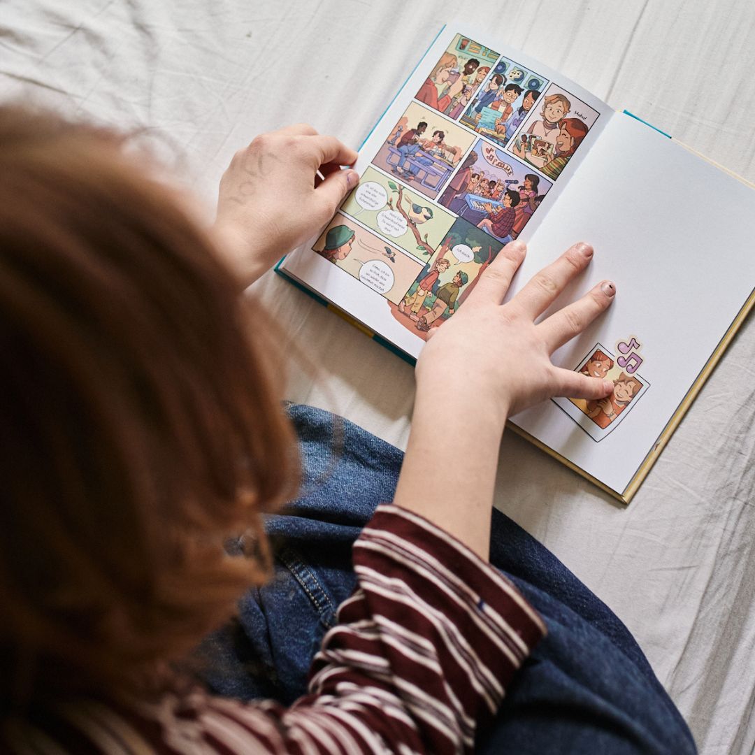 Ein Kind mit hellbraunem Haar und gestreiftem Hemd liest auf einem Bett mit weißen Laken den bunten Comic "Generation Glücklich" aus dem Zuckersüß Verlag und zieht, wie viele Kinder der Generation Glücklich, Geschichten dem Bildschirm vor.
