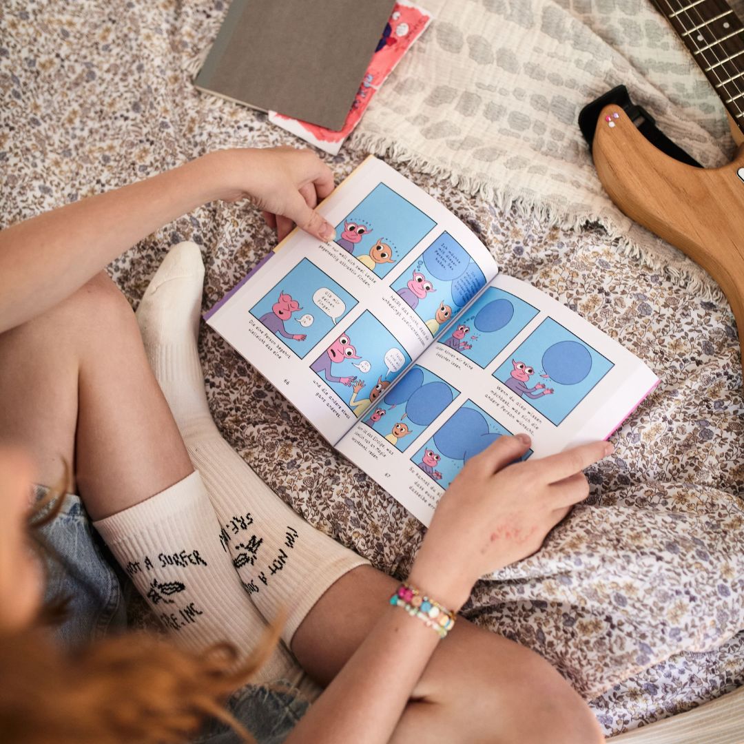 Eine Person sitzt im Schneidersitz auf einer gemusterten Tagesdecke und liest „How to love“ vom Zuckersüß Verlag – einen farbenfrohen Comic, der vielleicht eine willkommene Abwechslung zu den vielen Nachrichten über Dating oder das Navigieren durch Beziehungen bietet. Sie trägt weiße Socken mit schwarzer Aufschrift, ein Perlenarmband und neben ihr liegt eine Gitarre.