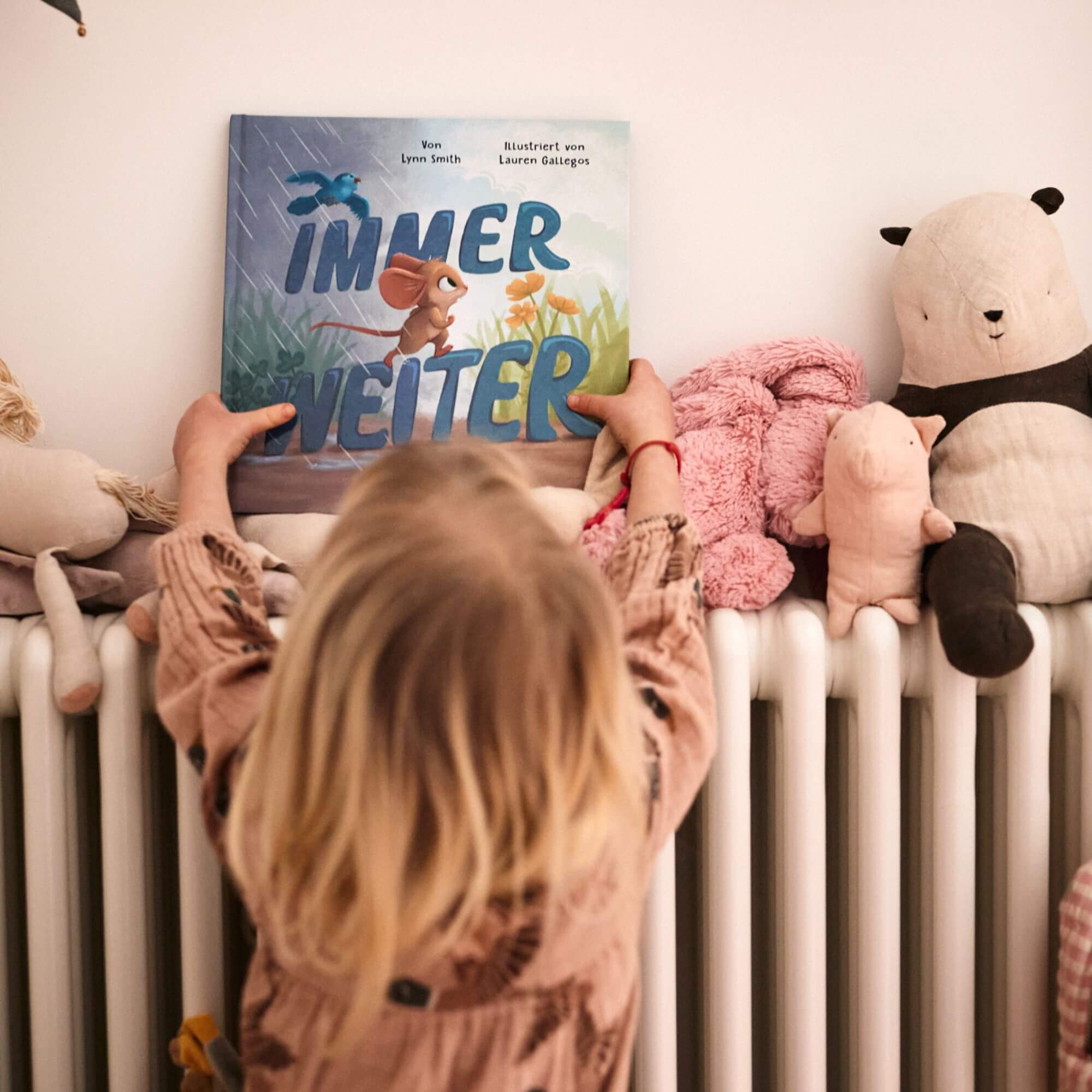 Ein kleines Kind mit blonden Haaren in einem hellbraunen Outfit hält Immer weiter vom Zuckersüß Verlag vor einer Reihe von Stofftieren hoch und erzählt eine Geschichte über Emotionen Kinder und wie man Gefühle bewältigen kann.