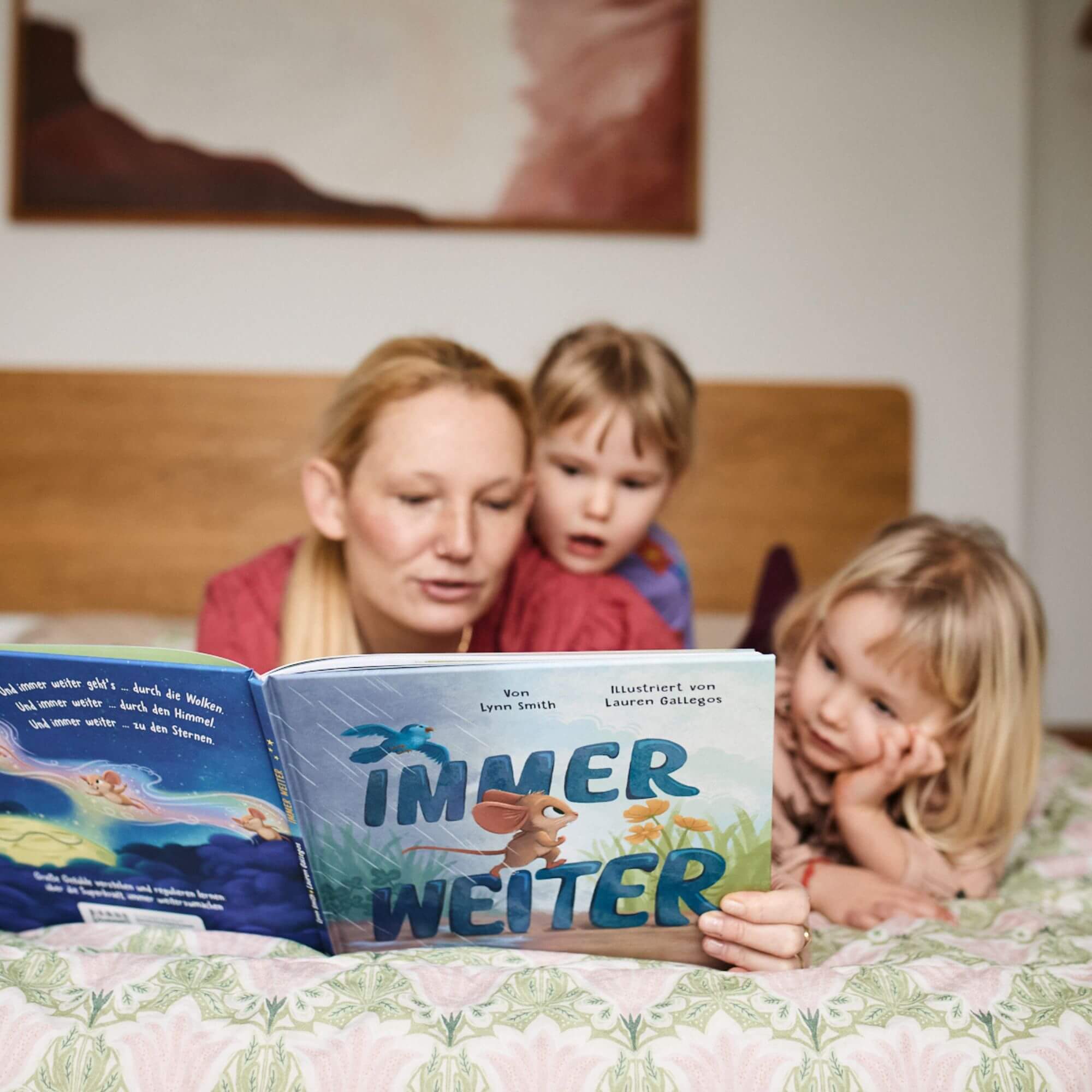 Eine Frau liest zwei kleinen Kindern auf einem Bett "Immer weiter" aus dem Zuckersüß Verlag vor; das eine Kind sieht aufgeregt aus, während das andere aufmerksam zuhört und etwas über den Umgang mit Emotionen und Gefühlen lernt.