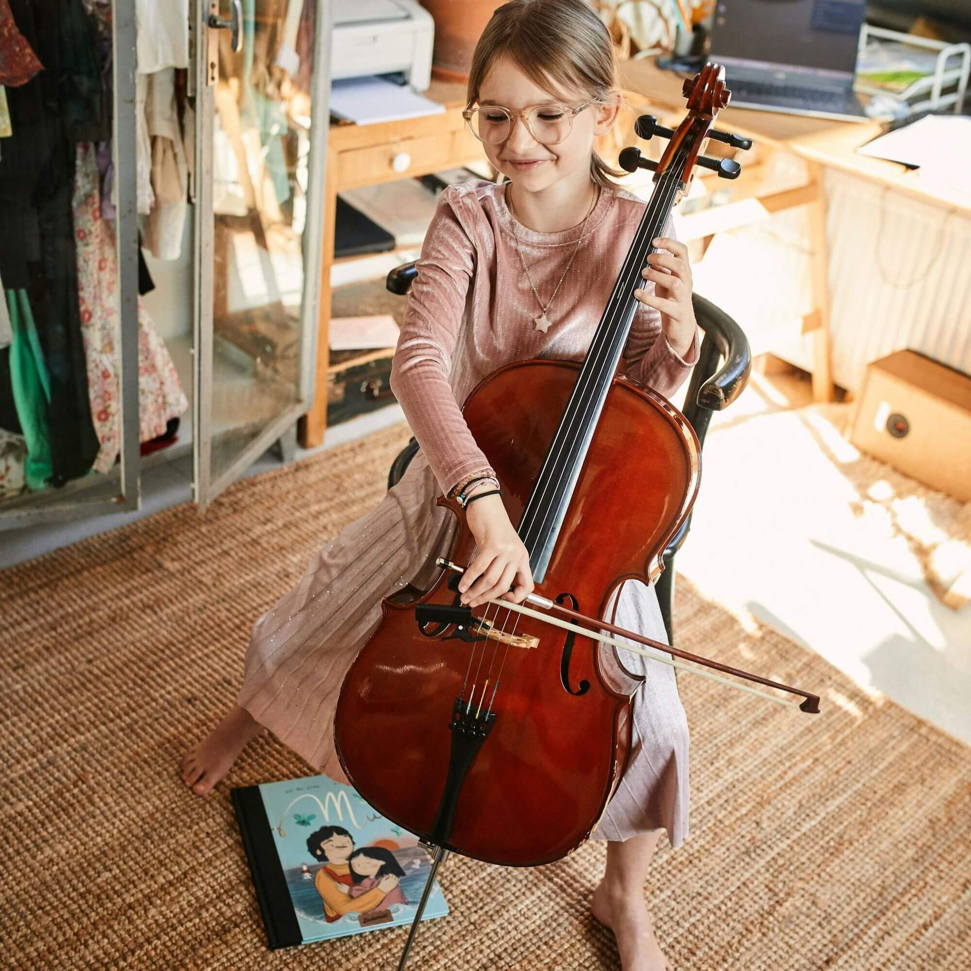 Ein junges Kind mit Brille und rosa Kleid spielt lächelnd und barfuß in einem gemütlichen Zimmer Cello. Neben ihr auf dem gewebten Teppich liegt "Mut", ein Kinderbuch von Wai Mei Wong, dessen illustrierter Einband geschlossen ist, während sie sich auf einen Stuhl setzt.