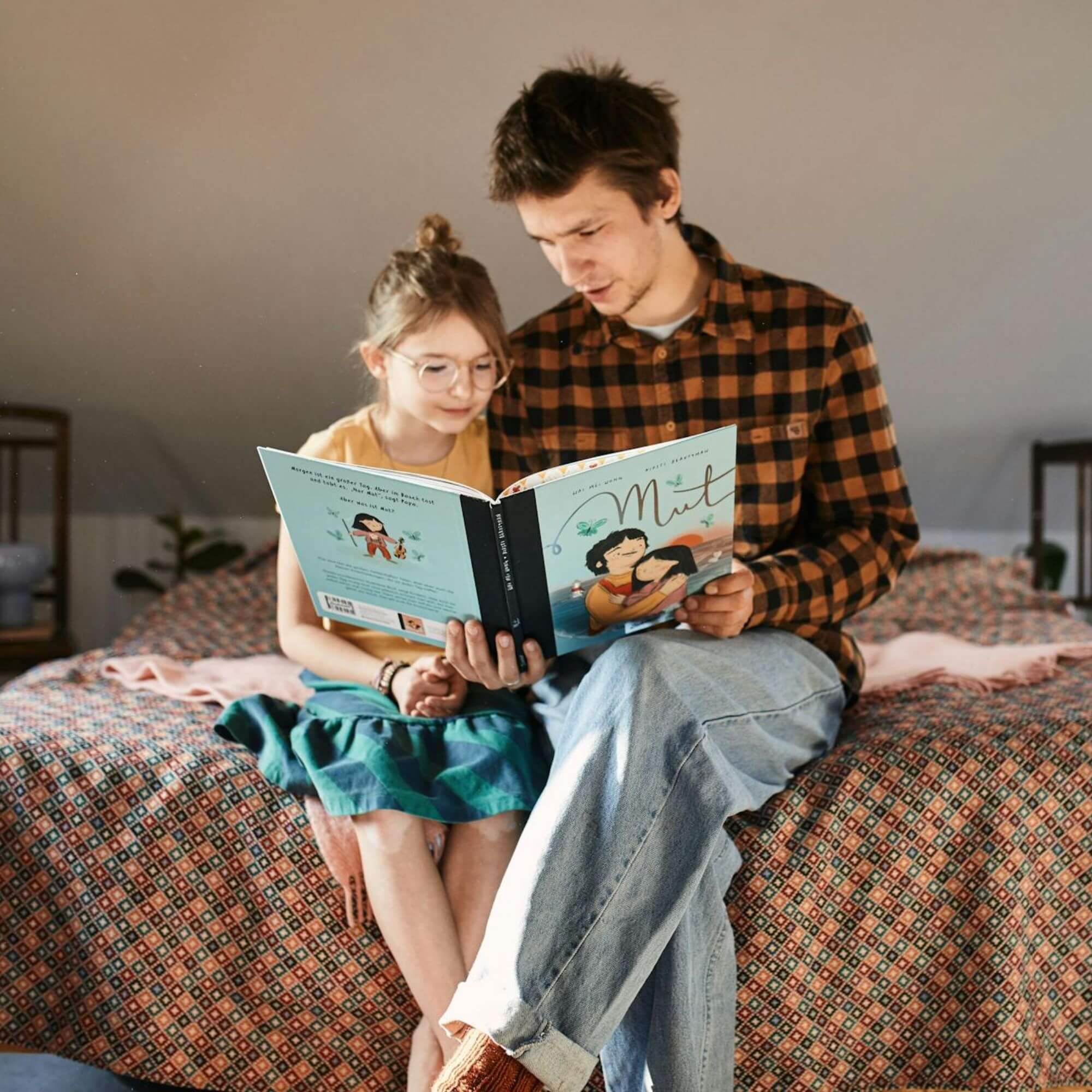 Eine erwachsene Person und ein Kind sitzen auf einer gemusterten Bettdecke und lesen "Mut" von Wai Mei Wong. Die erwachsene Person trägt ein kariertes Hemd und Jeans, das Kind eine Brille und einen grünen Rock. Sie verbringen einen ruhigen Moment miteinander, beide konzentriert auf das inspirierende Buch.