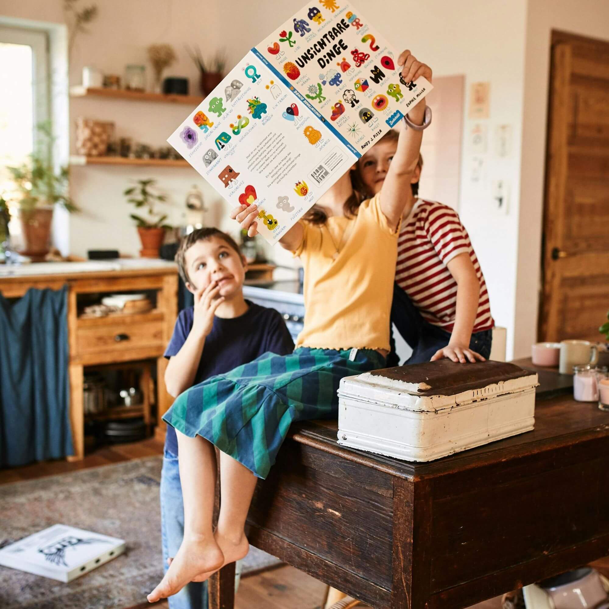 Zwei Kinder sitzen an einem Holztisch in einem gemütlichen Raum und halten gemeinsam das Bilderbuch "Unsichtbare Dinge" von Andy J. Pizza und Sophie Miller hoch. Eines zeigt begeistert auf eine Stelle im Buch, während das andere das Buch über den Kopf hält.