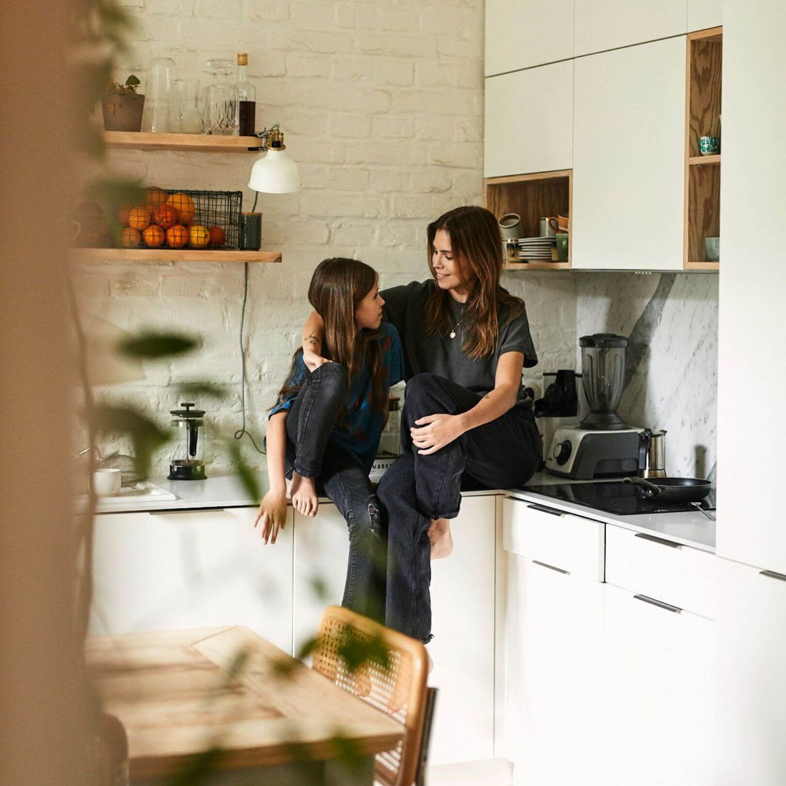 Eine Frau und ein junges Mädchen sitzen an einer Küchentheke und lächeln sich in einem hellen, modernen Raum an. Die Szene zeigt das "Young Feminist Starter Pack" des Zuckersüß Verlags inmitten von weißen Schränken, Holzregalen, Obst und Küchengeräten.