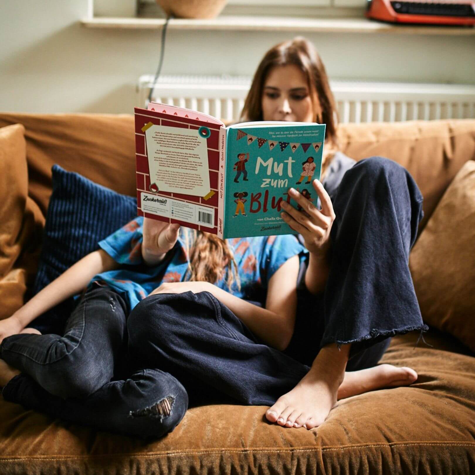 Zwei Menschen entspannen sich auf einem braunen Sofa und lesen "Mut zum Blut". Das Young Feminist Starter Pack des Zuckersüß Verlags hilft Kindern in der Pubertät dabei, sich selbst besser kennenzulernen.