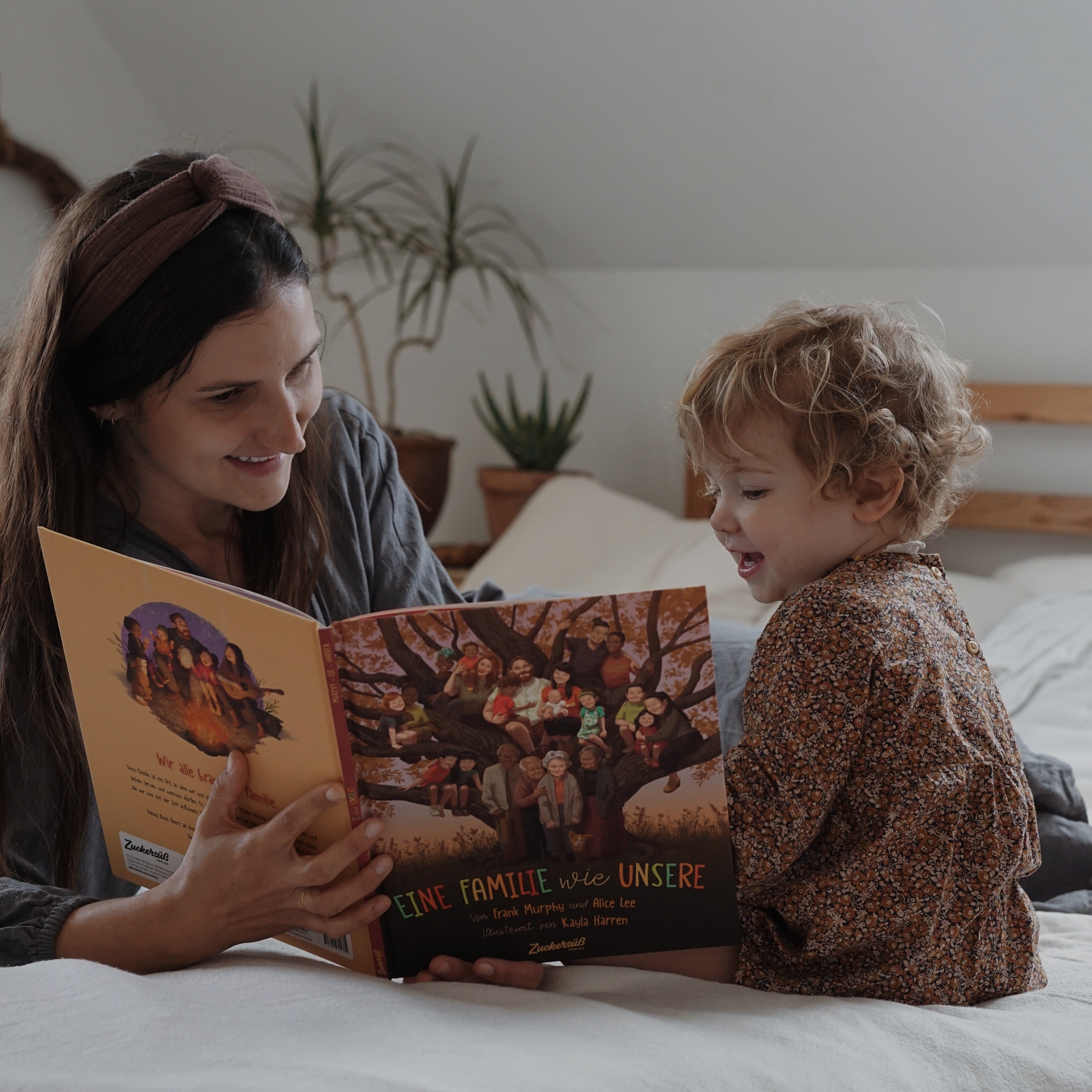 Eine lächelnde erwachsene Person mit Kind sitzt auf einem Bett und liest "Das Richtig-Gute-Nacht-Paket" vom Zuckersüß Verlag. Der sanft beleuchtete Raum mit einer Pflanze und Holzmöbeln strahlt Wärme und Geborgenheit aus.