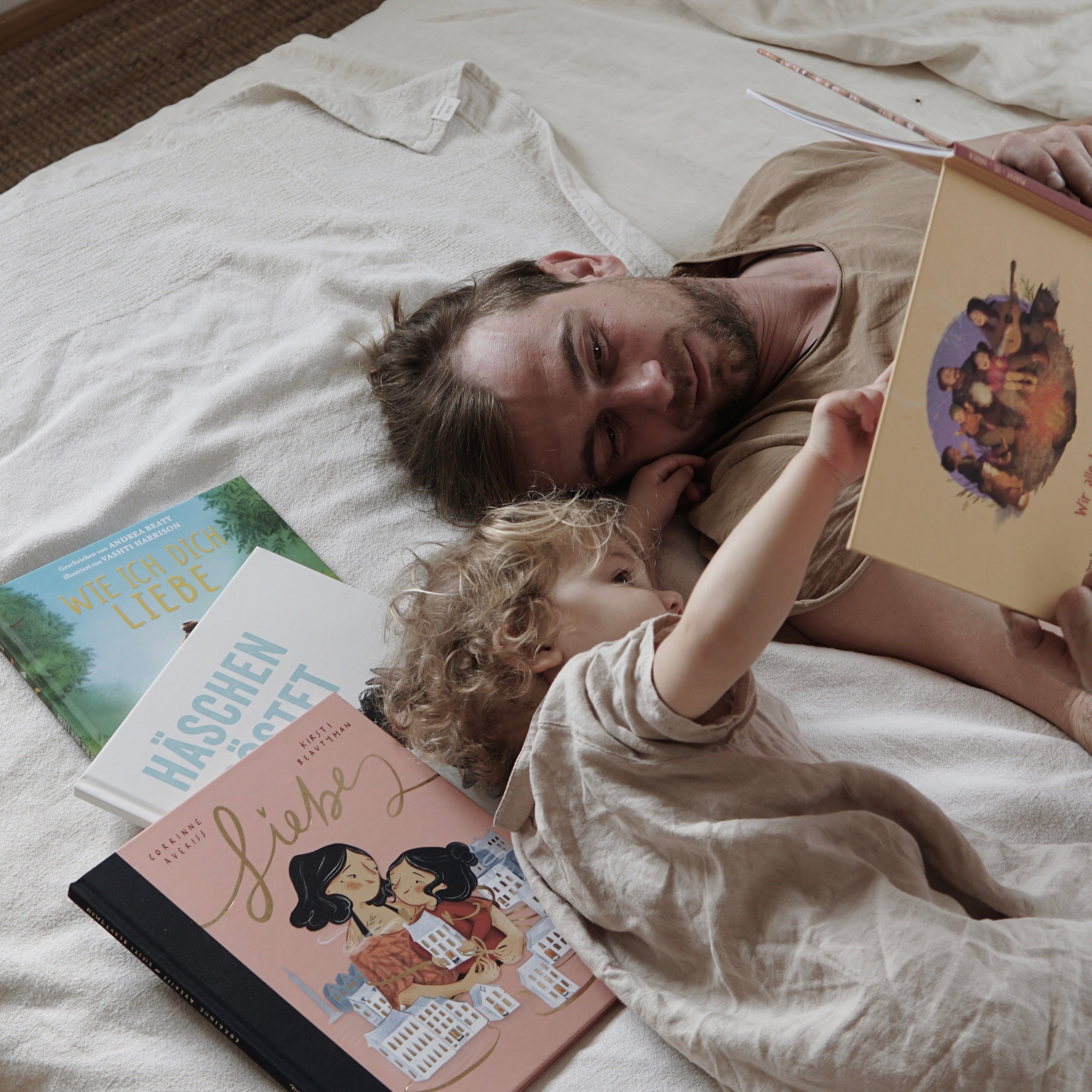 Ein Mann und ein Kind liegen auf einem Bett und schauen sich gemeinsam ein Kinderbuch an. Um sie herum liegen weitere Titel aus dem „Richtig-Gute-Nacht-Paket“ des Zuckersüß Verlags.