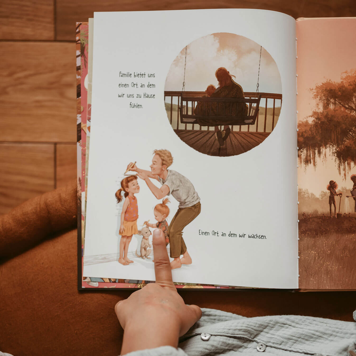 Eine Kinderhand zeigt auf eine aufgeschlagene Seite aus dem „Welt verändern Starter Pack“ des Zuckersüß Verlags. Die Doppelseite zeigt liebevolle Familienszenen: oben eine Bezugsperson mit Kind auf einer Holzschaukel im Sonnenuntergang, unten ein Elternteil mit zwei Kindern und einem Hund. Der Text spricht über Familie als Ort des Zuhauseseins und Wachsens.
