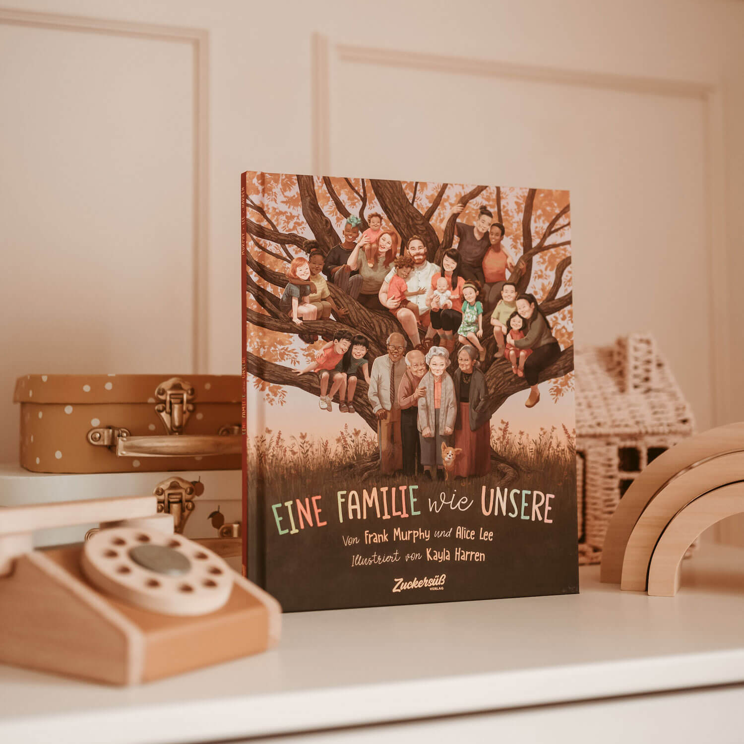 Auf einem Holzregal steht das Bilderbuch "Eine Familie wie unsere" von Frank Murphy und Alice Lee, das eine vielfältige Mehrgenerationenfamilie um einen Baum herum zeigt. Daneben stehen Holzspielzeug und Korbwaren.