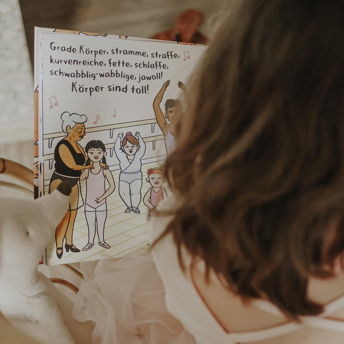 Ein Kind in einem rosafarbenen Kleid hält ein aufgeschlagenes "Körper sind toll"-Buch von Tyler Feder in der Hand, das verschiedene Balletttänzerinnen und -tänzer und einen Text zeigt, der alle Körpertypen und Körperwahrnehmung feiert.