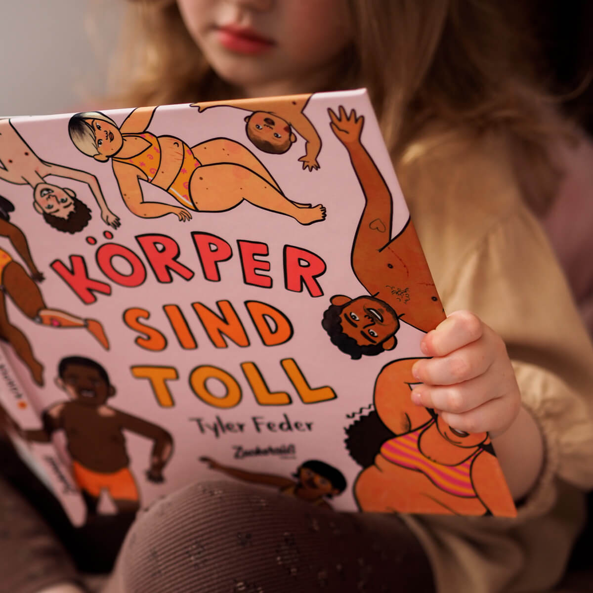 Ein kleines Kind liest "Körper sind toll" von Tyler Feder, ein Buch mit verschiedenen Körpern auf dem Cover, das verschiedene Körpertypen und eine positive Körperwahrnehmung feiert.