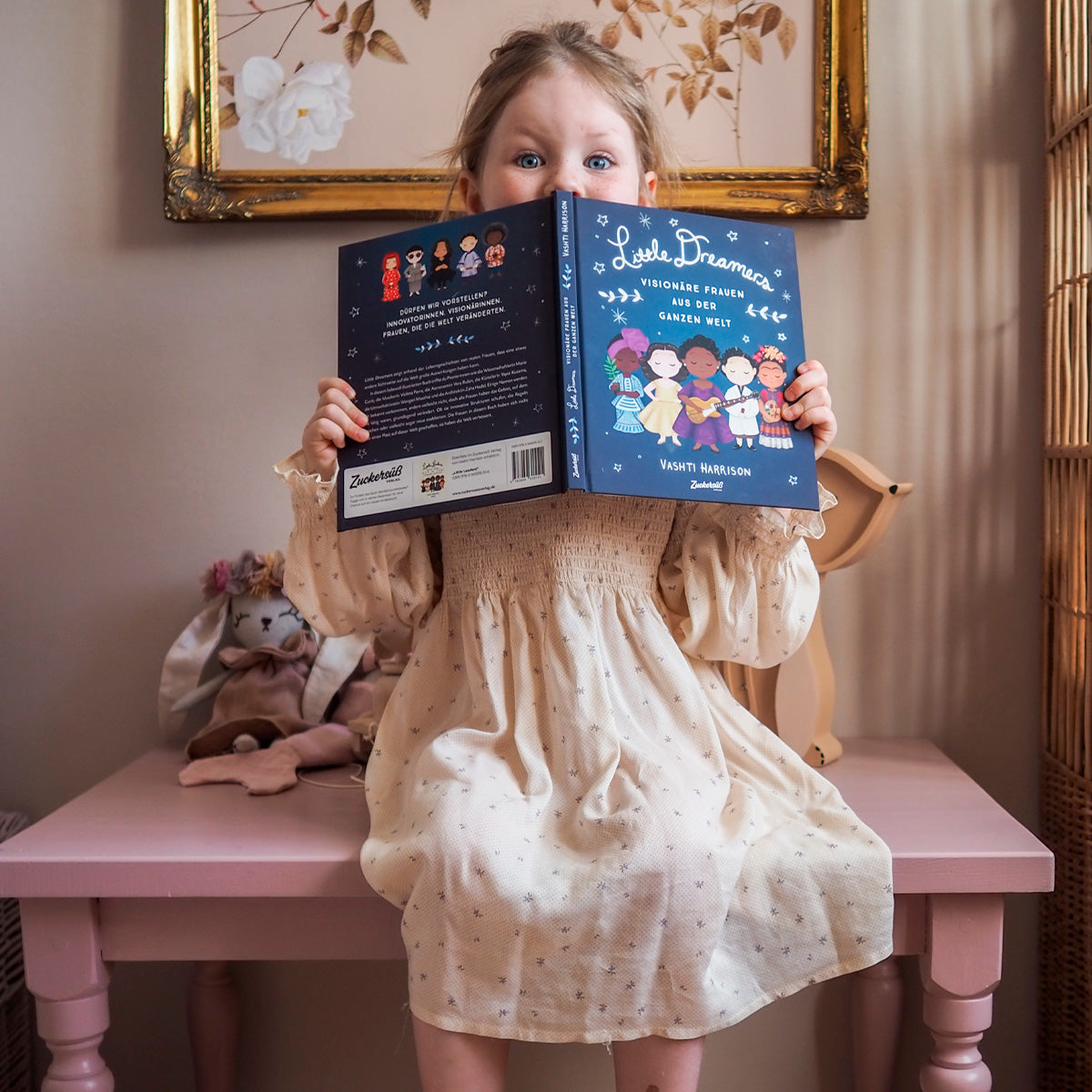 Ein junges Kind in einem cremefarbenen Kleid sitzt an einem rosa Tisch und liest „Little Dreamers“ von Vashti Harrison. Hinter ihr stehen ein Plüschhase und ein gerahmtes Blumenbild.