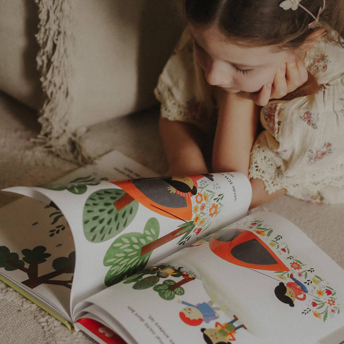 Ein Kind mit hellbraunen Haaren und geblümtem Kleid liegt auf einem Teppich und blättert im Bilderbuch „Manchmal bin ich alles, was ich brauche“ von Juliana Perdomo. Aufgeschlagen ist eine bunte Illustration mit Bäumen, Blumen und Figuren in orangefarbenen Umhängen.