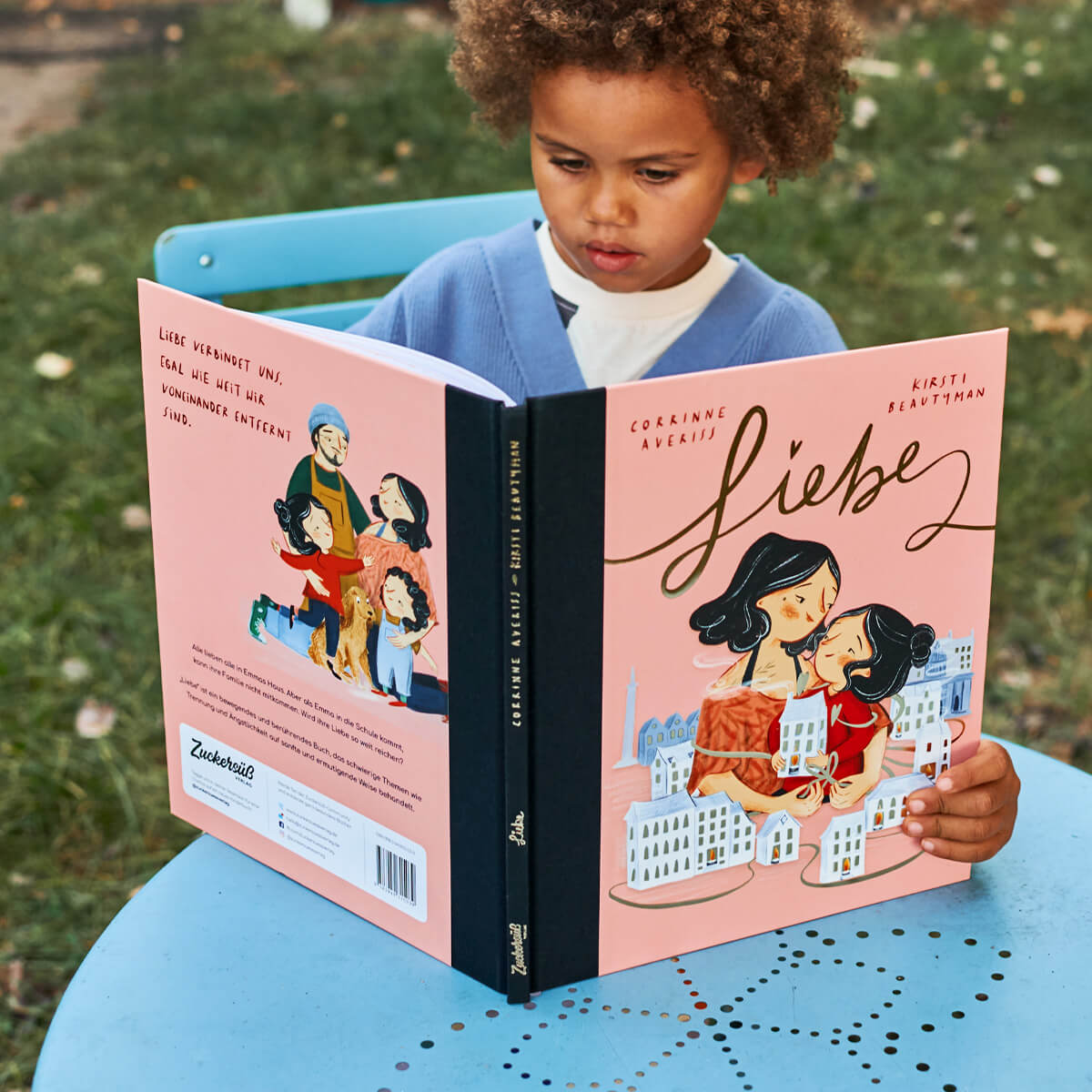Ein Kind mit lockigem Haar sitzt an einem blauen Tisch im Freien und liest das Buch „Liebe“ von Corrinne Averiss aus dem Paket „Mut & Liebe“ des Zuckersüß Verlags. Das Cover zeigt eine innige Umarmung zwischen zwei Personen, umgeben von stilisierten Häusern auf rosafarbenem Hintergrund – eine liebevoll illustrierte Szene voller Wärme und Geborgenheit.