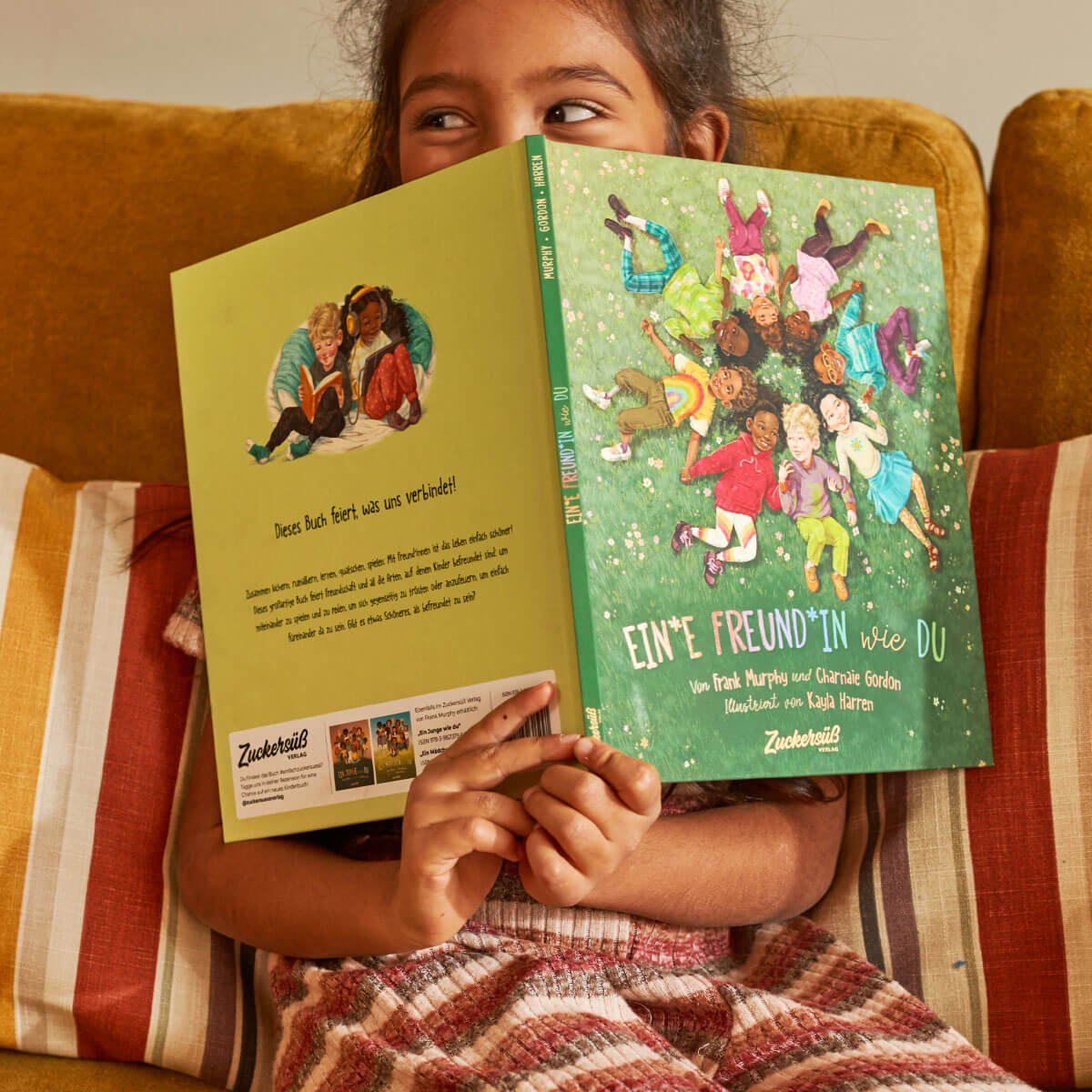 Ein lächelndes Kind auf einer goldenen Couch hält "Ein*e Freund*in wie du" von Frank Murphy & Charnaie Gordon hoch, wobei es sein Gesicht teilweise verdeckt. Das Cover des Buches zeigt illustrierte Kinder, die auf einer Wiese die Freundschaft feiern.