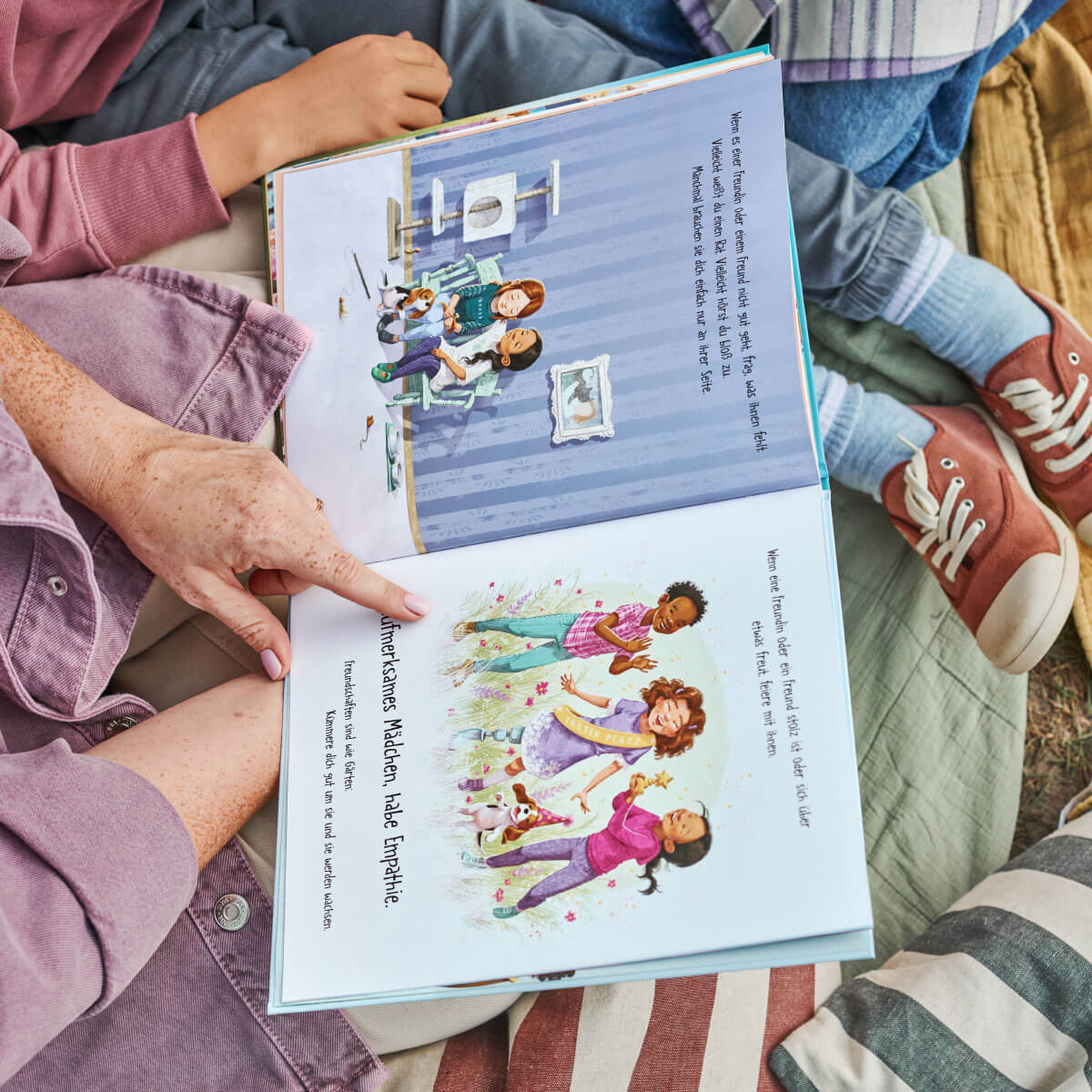 Eine erwachsene Person liest einem Kind, das auf einer gestreiften Decke im Freien sitzt, das Kinderbuch „Ein Mädchen wie du“ von Frank & Carla Murphy vor. Die aufgeschlagenen Seiten zeigen farbenfrohe Illustrationen von Kindern mit Hunden und vermitteln eine Botschaft über Freundschaft und Empathie.