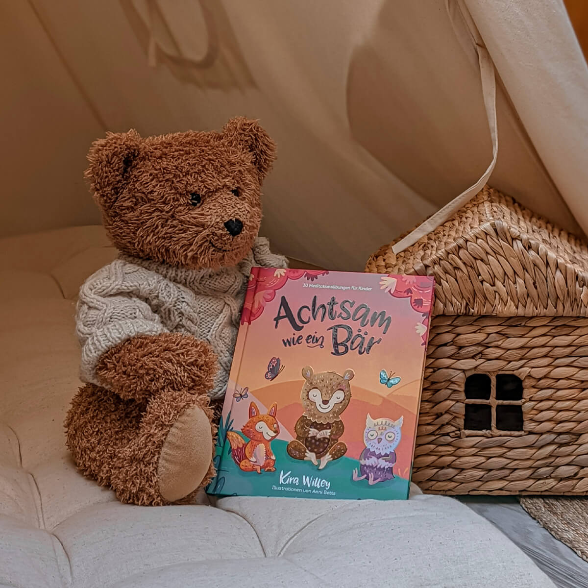 Ein brauner Teddybär im Pullover sitzt neben einem gewebten Spielzeughaus und hält "Achtsam wie ein Bär" von Kira Willey in der Hand, ein Kinderbuch mit Tierillustrationen und Meditationsübungen zum Stressabbau in der Familie.
